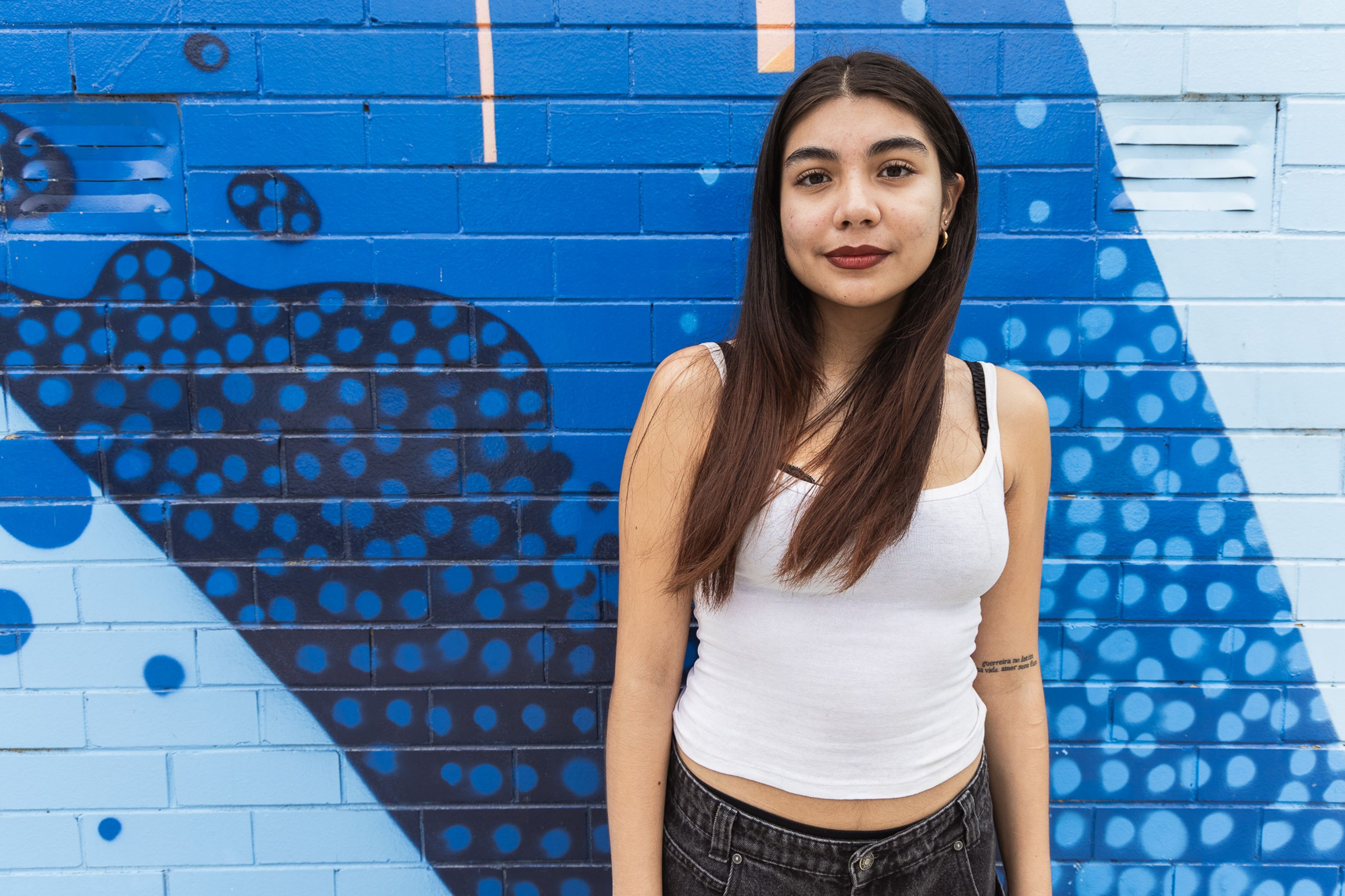 Val, a young woman with long dark hair, stands in front of vibrant blue brick wall.