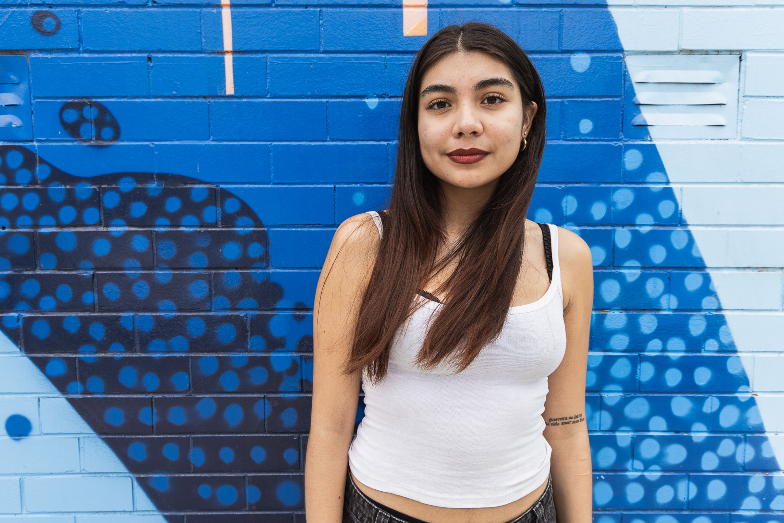 Val, a young woman with long dark hair, stands in front of vibrant blue brick wall.