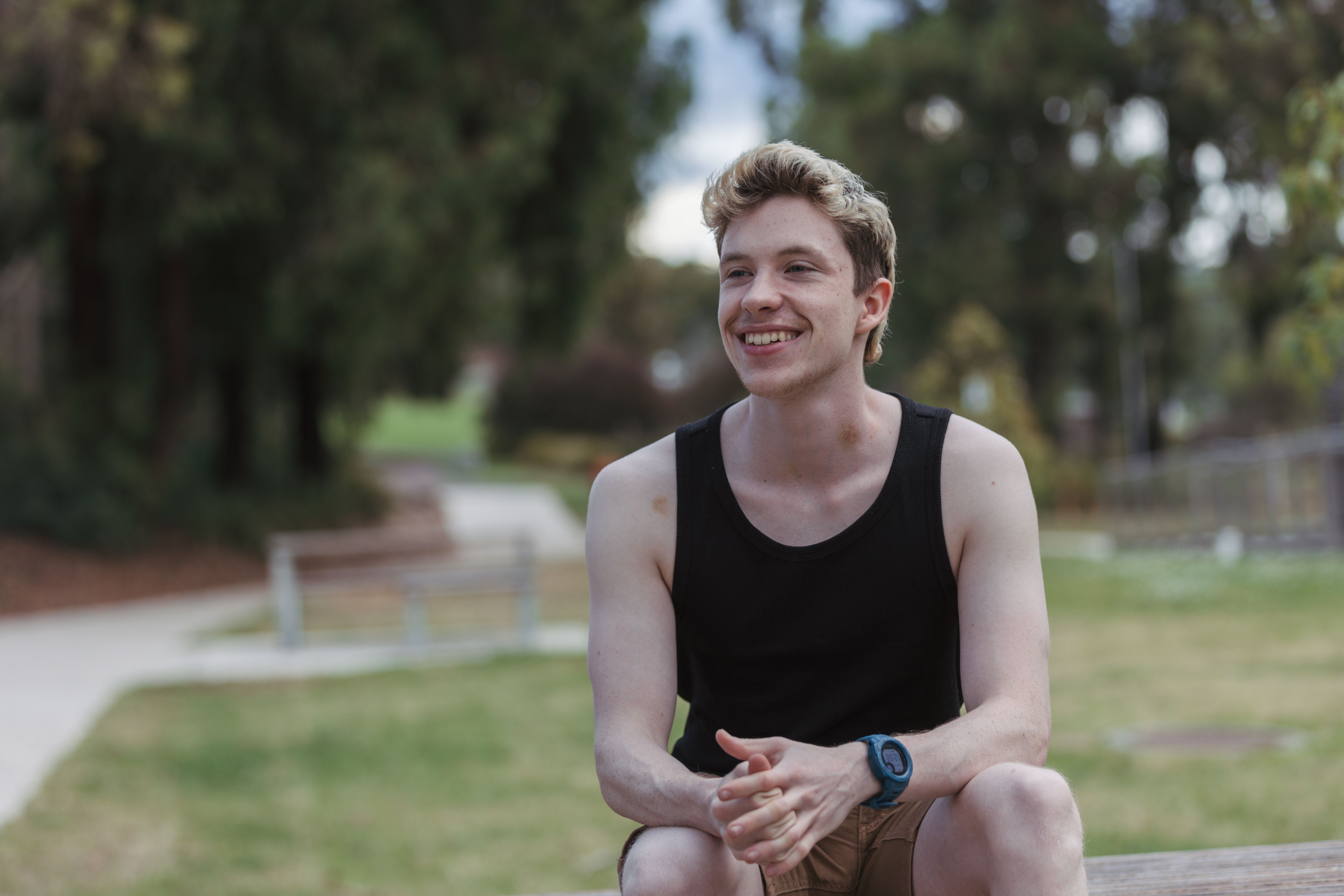 Young man with a black singlet sitting on a park table with hands in lap smiling and looking into the distance