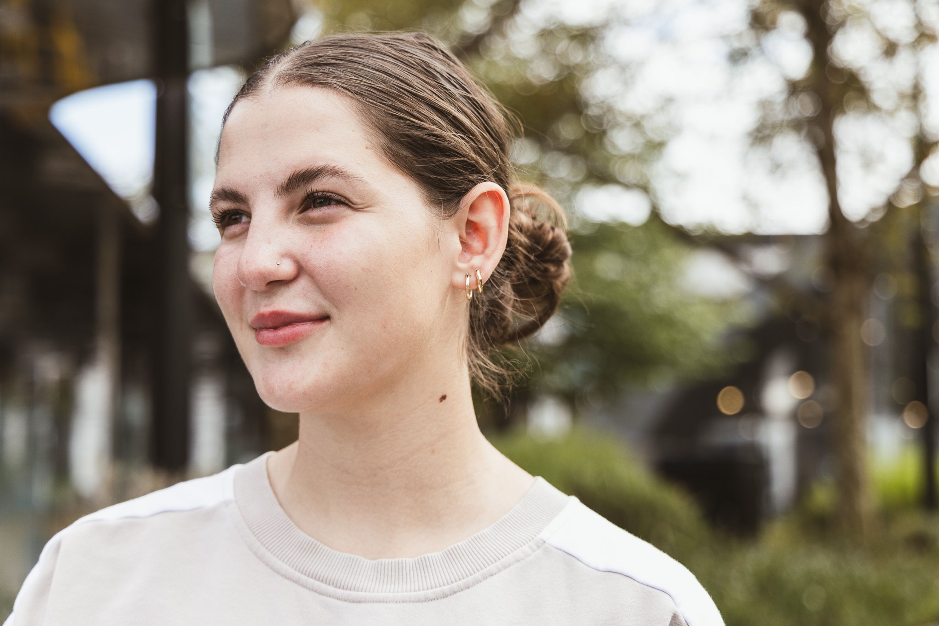 Abbey, a young woman with light skin and dark brown hair stands outdoors, looking to the side with a gentle smile.