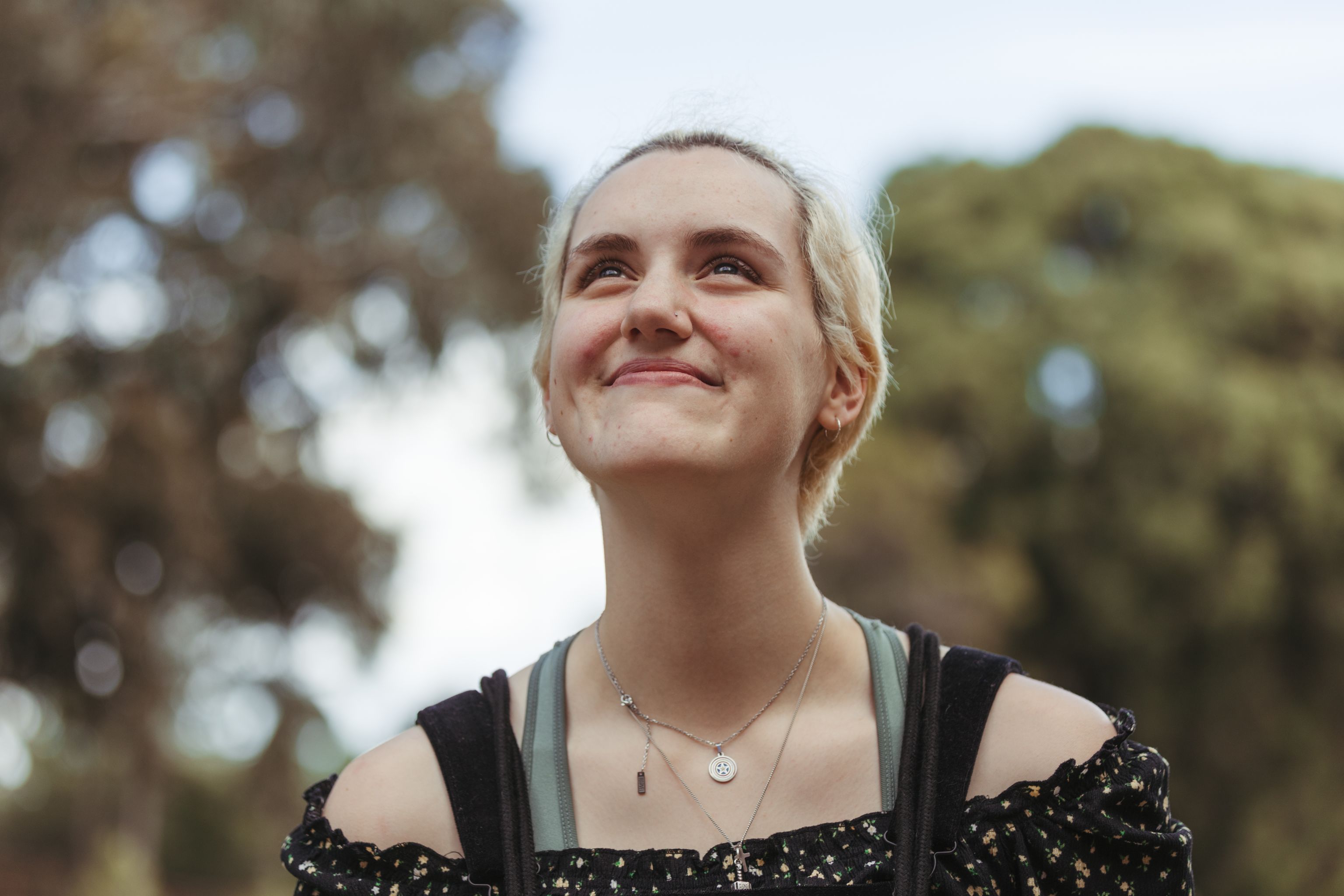 Young individual with short blond hair looking upwards and smiling, with trees in the background