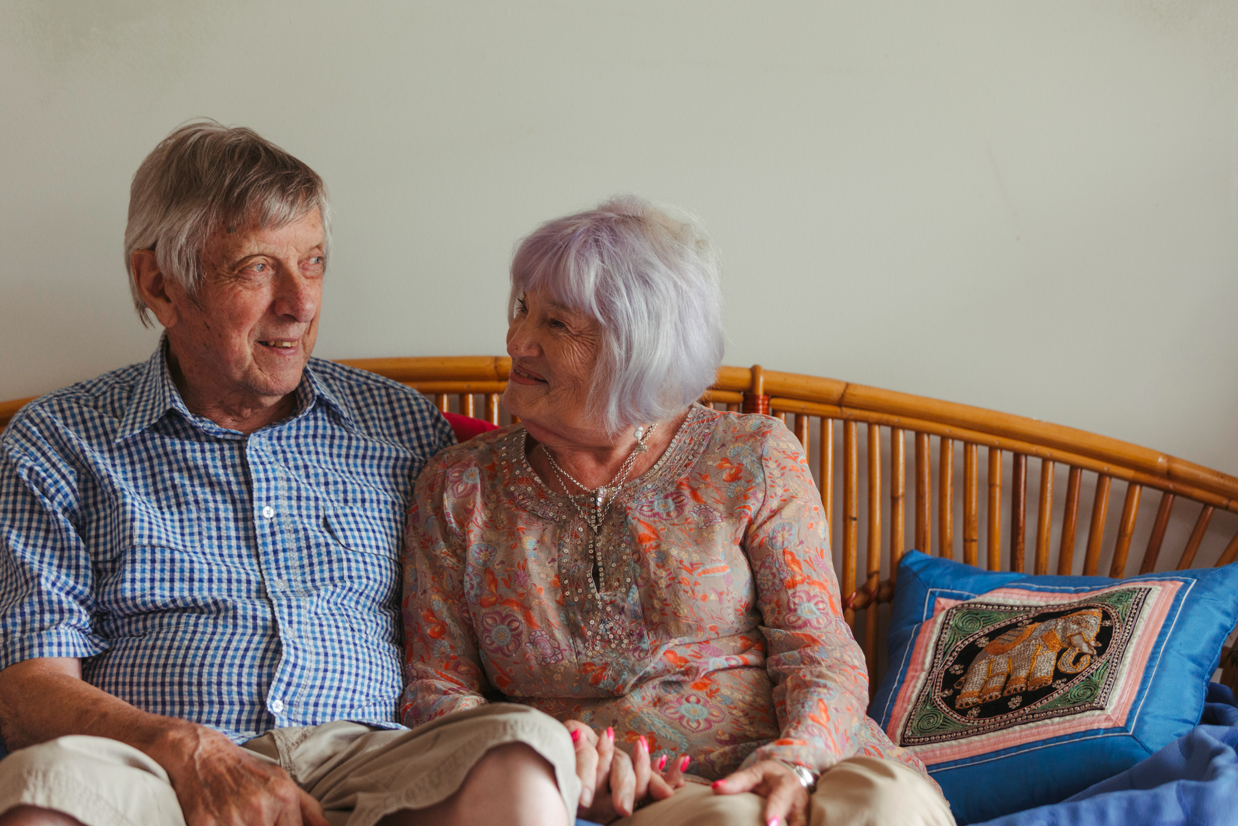 An older couple sits closely on a rattan couch, smiling warmly at each other in a cozy setting.