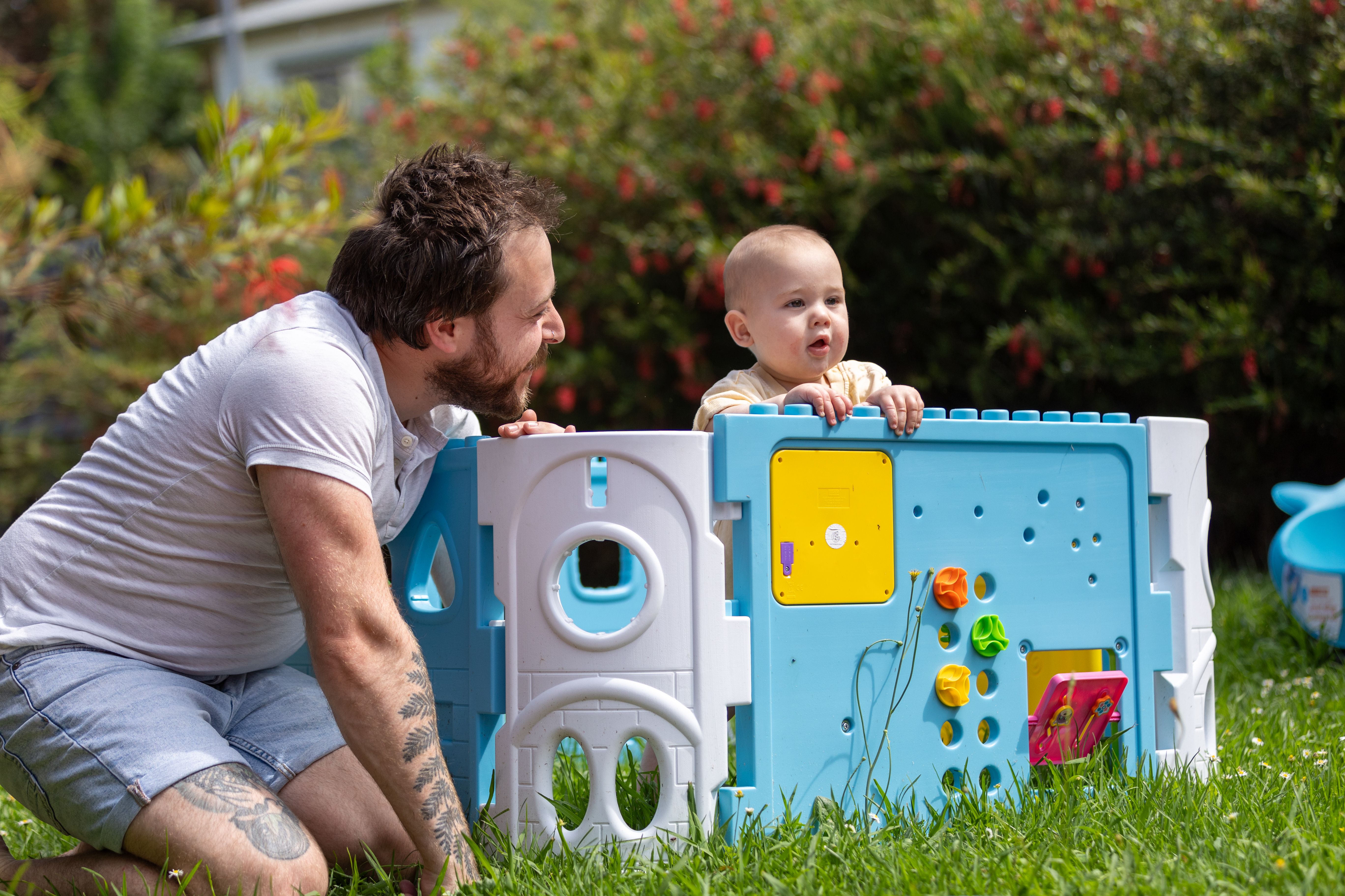 Young man in a garden kneeling next to his son who is playing in a toy enclosure
