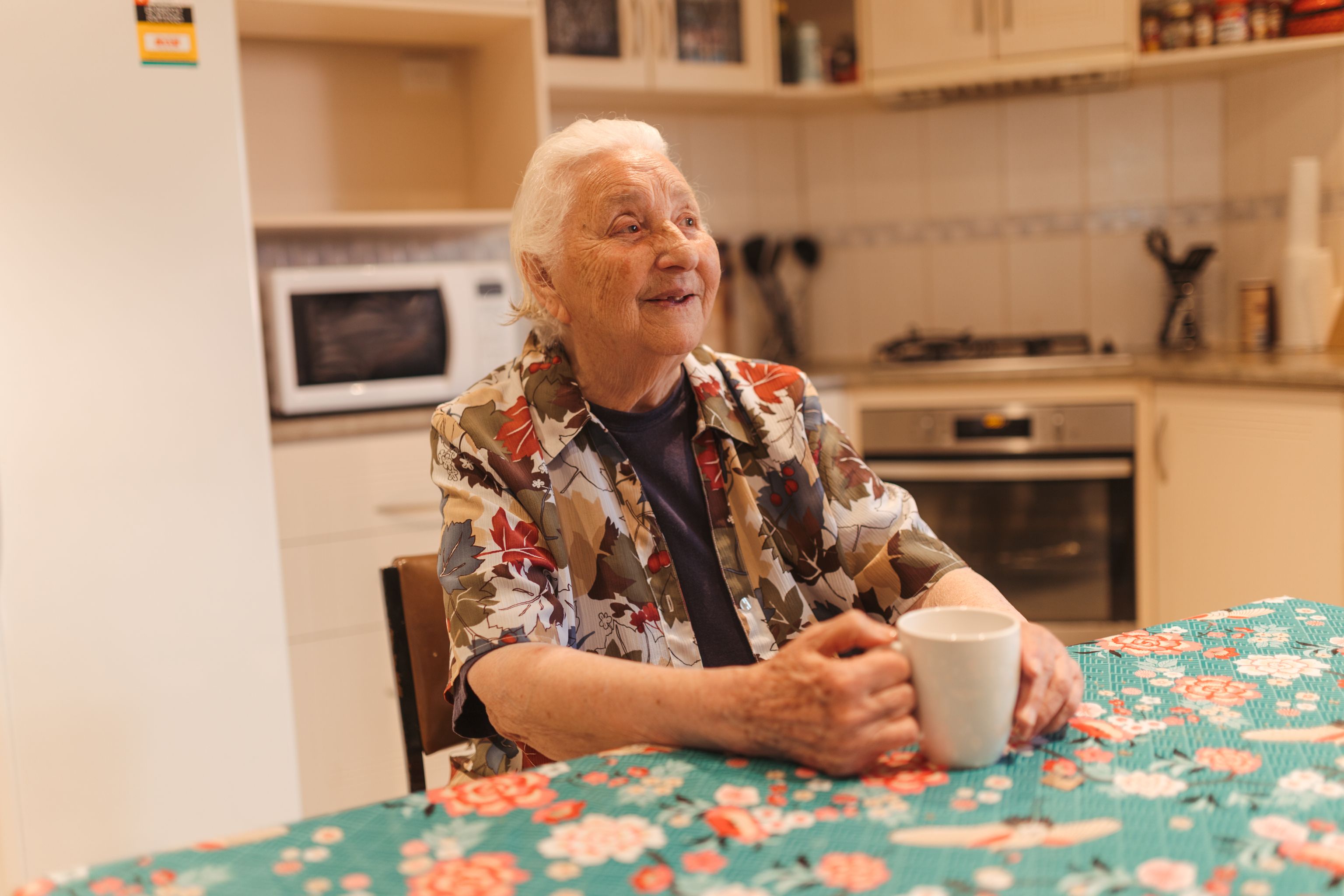 Elida, an 85-year-old woman, sits at a bright floral table in her kitchen. She’s holding a cup of tea and looking ahead.