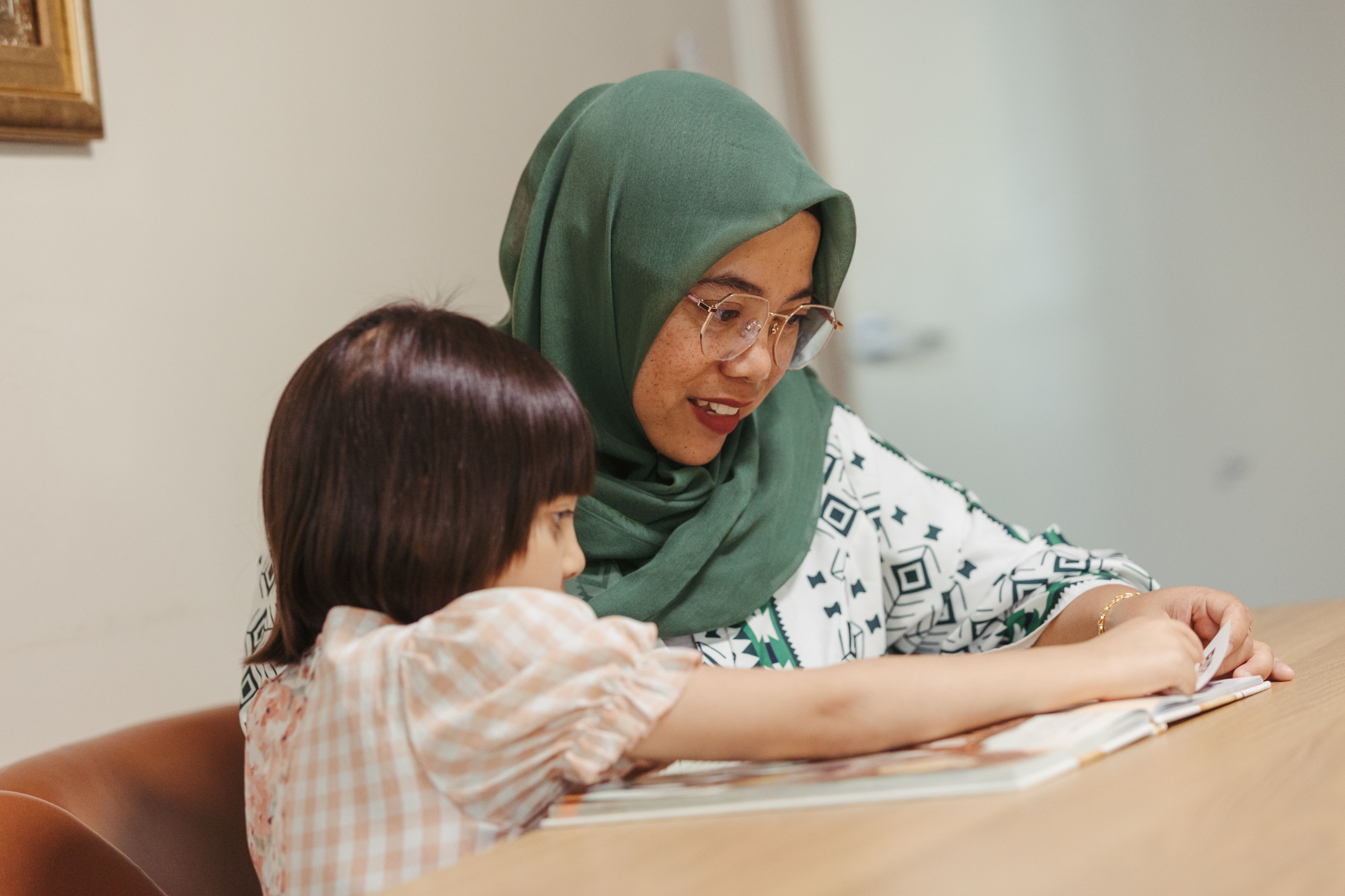 Women wearing a hijab sits at a table reading a book to a young girl