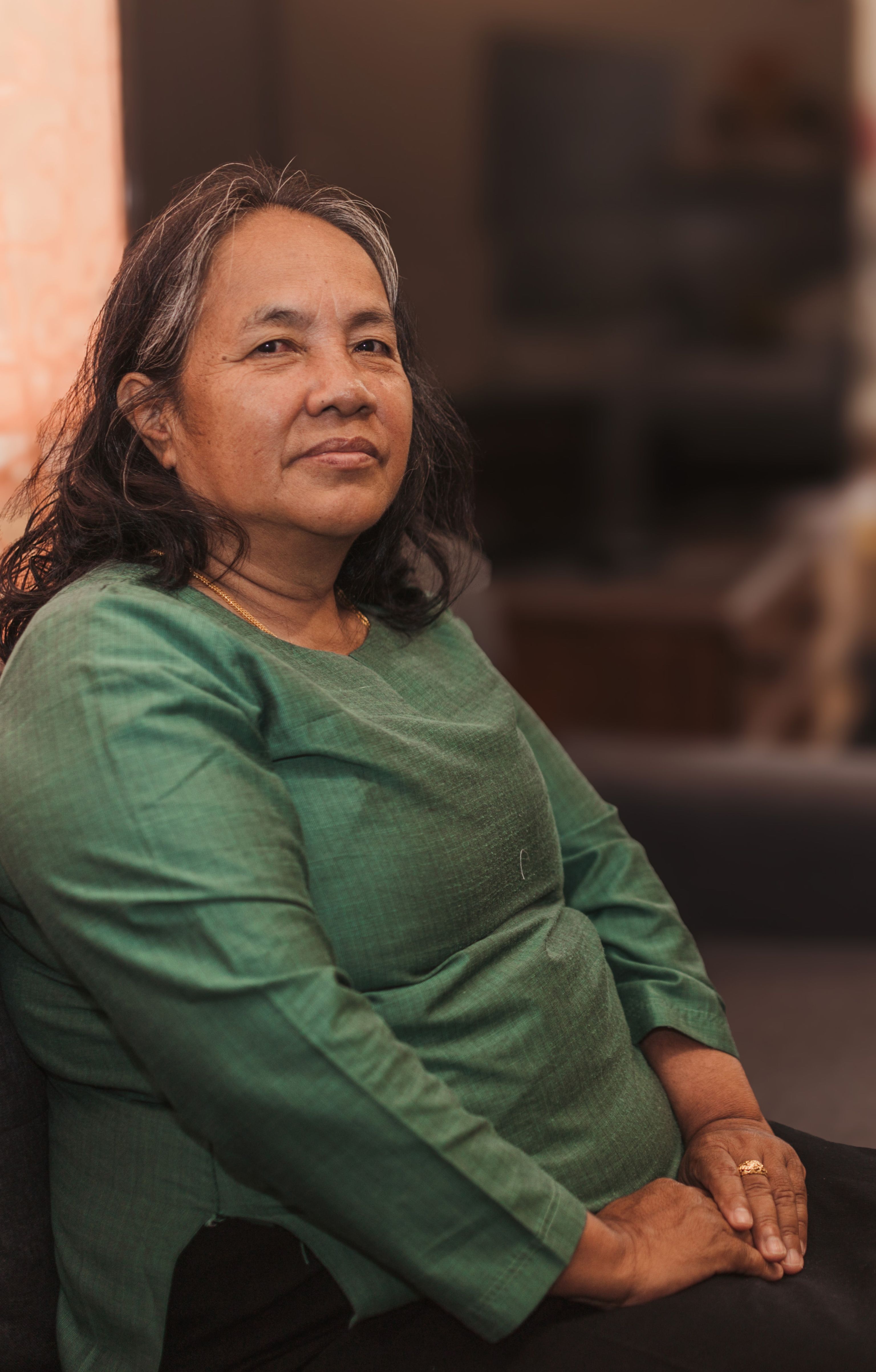 Mother Fung sits on a chair in her home with her hands on her lap. She looks at the camera with a content expression.