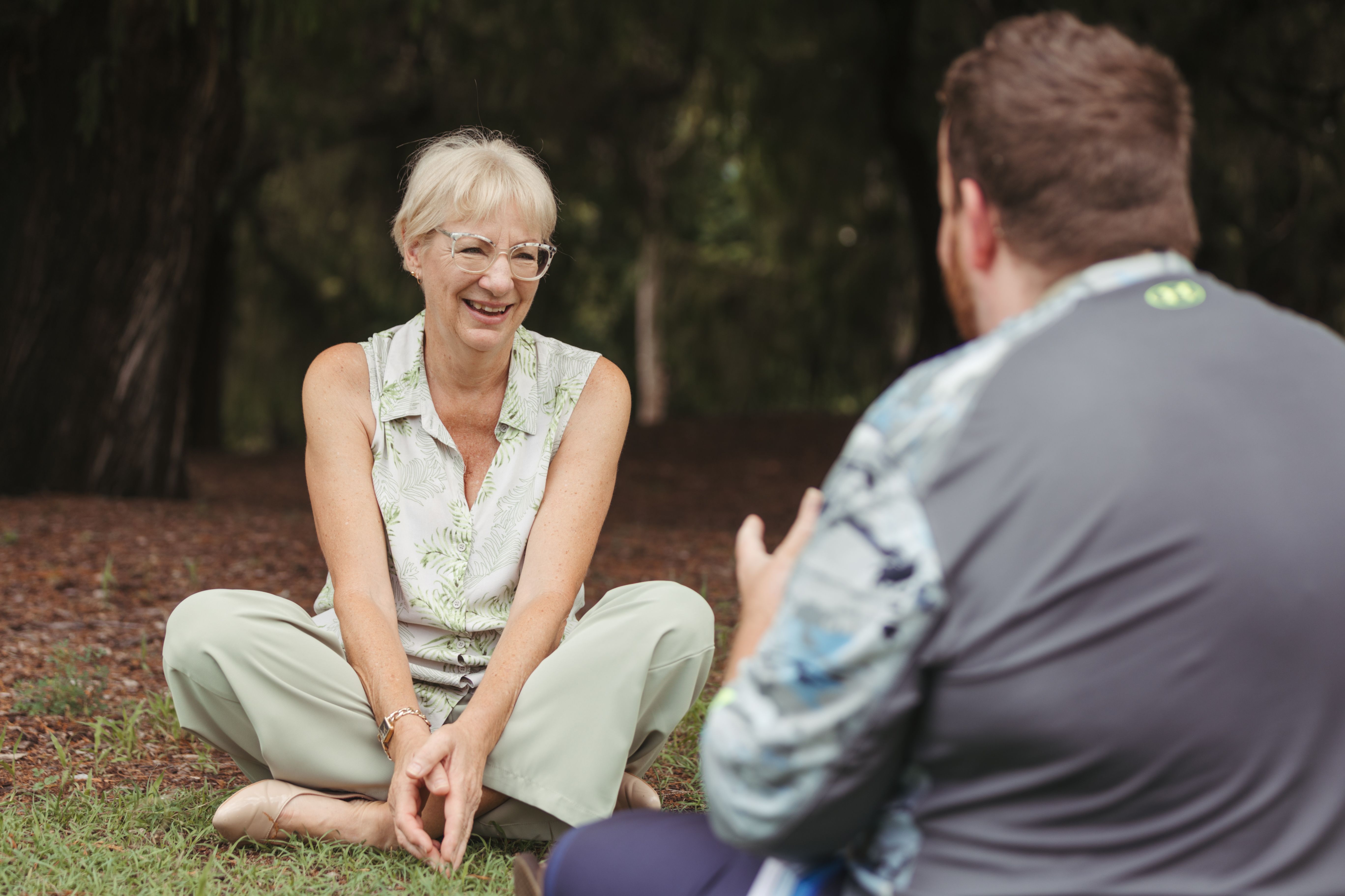 A middle aged women sitting crossed legged in a park chatting to a man sitting opposite her