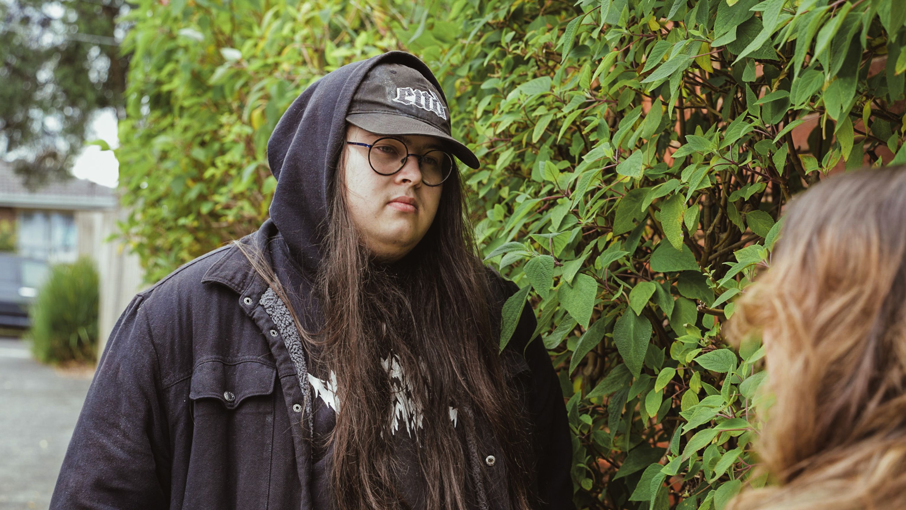 Young adult Ethan is outside, wearing a black hoodie and a hat. He stands next to a wall covered in green plants and is looking at a person we can't see.