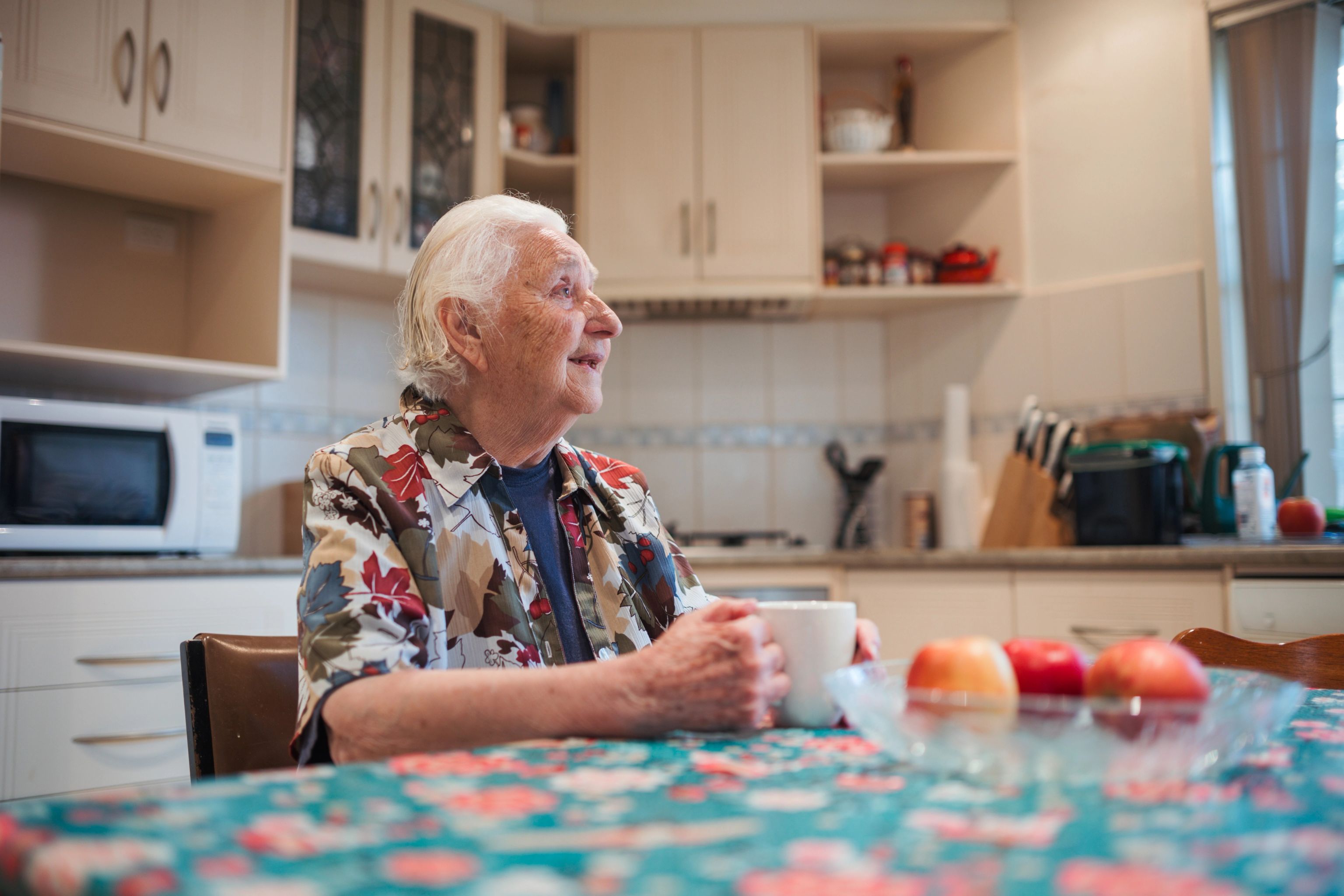 A woman sits at her kitchen table, holding a mug. She's smiling and looking away from the camera.