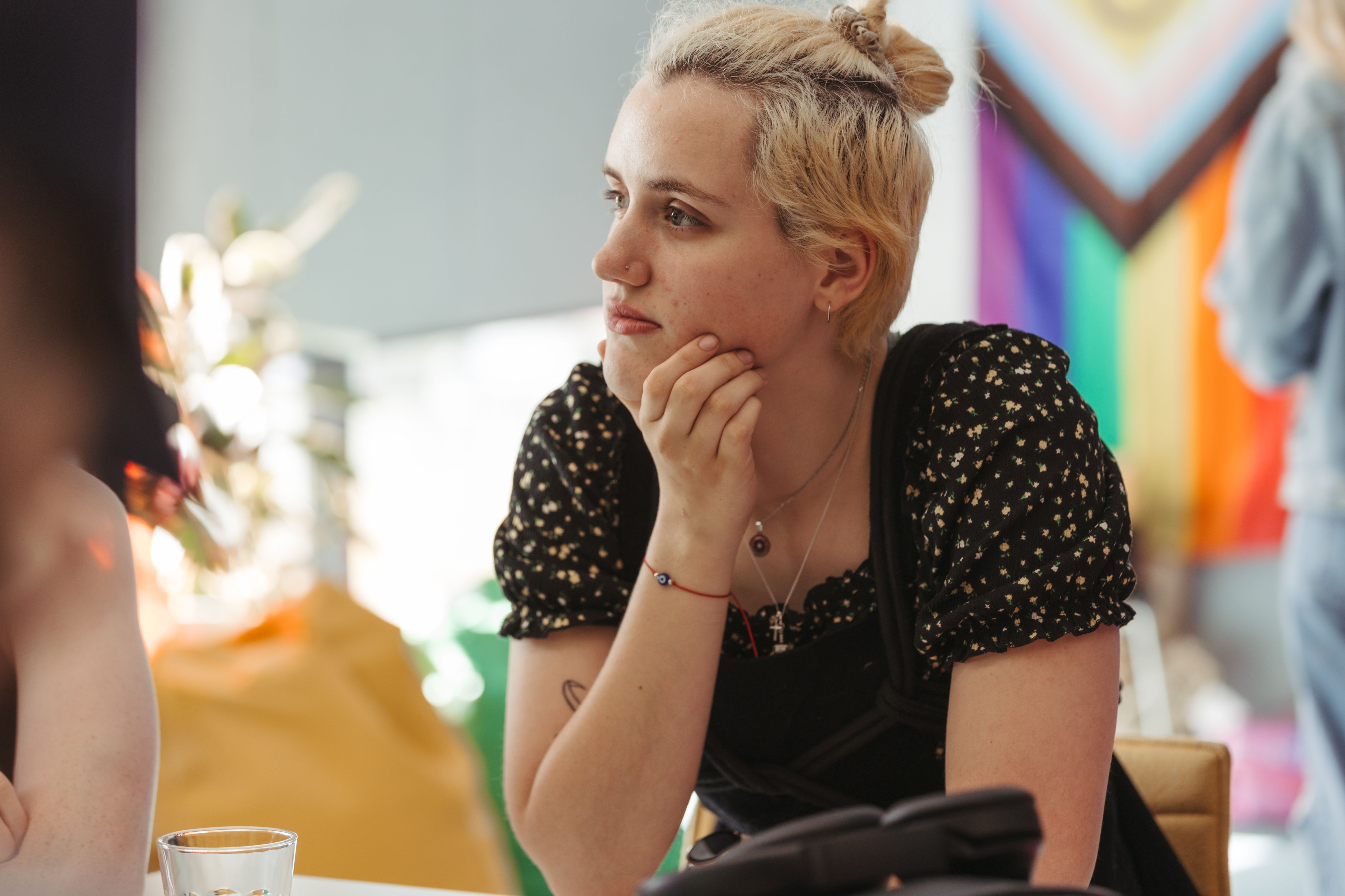 Young woman sitting at a table with her chin resting on her hand and a pride flag in the background