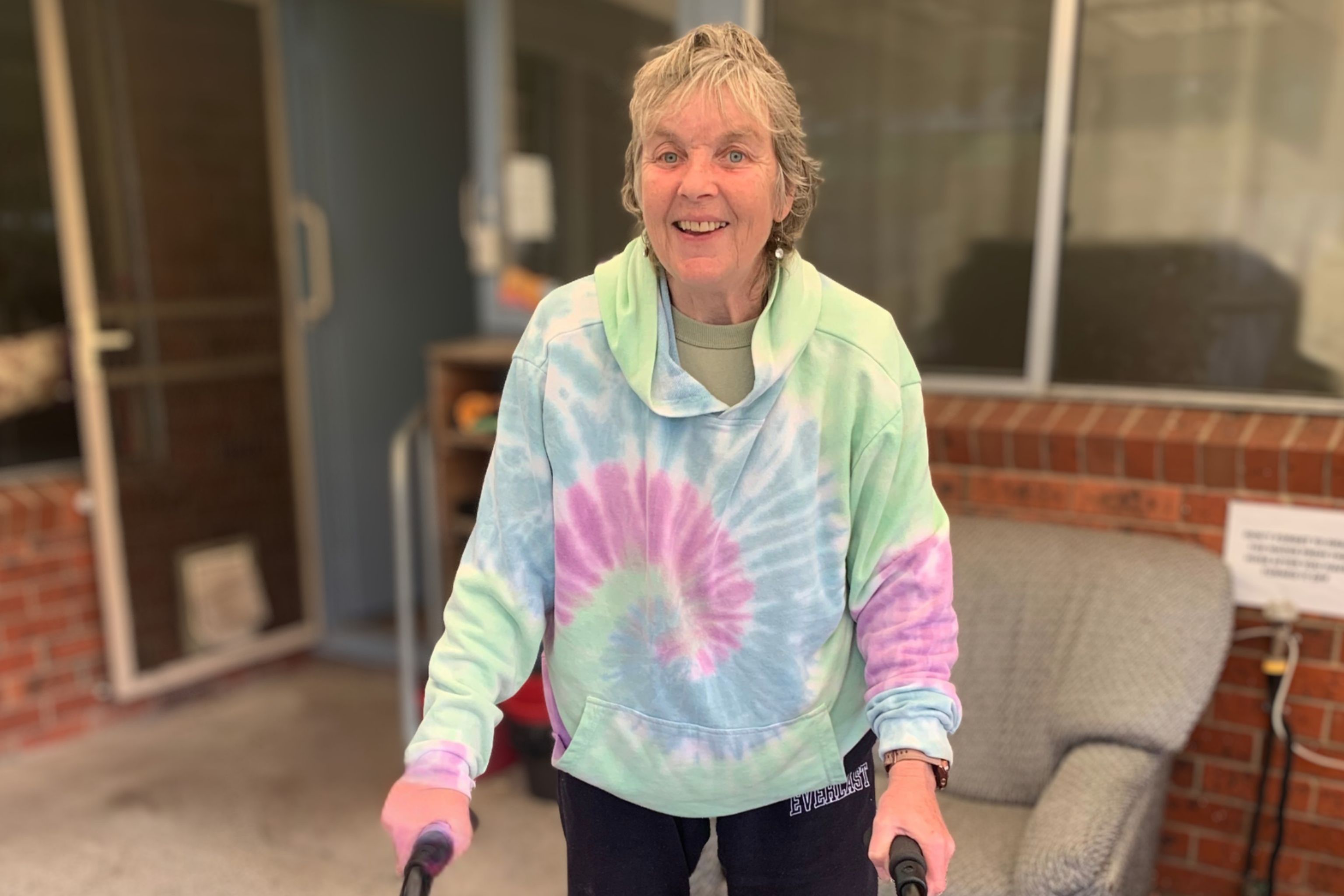 Person standing indoors using a walking aid, wearing a colourful tie-dye hoodie.