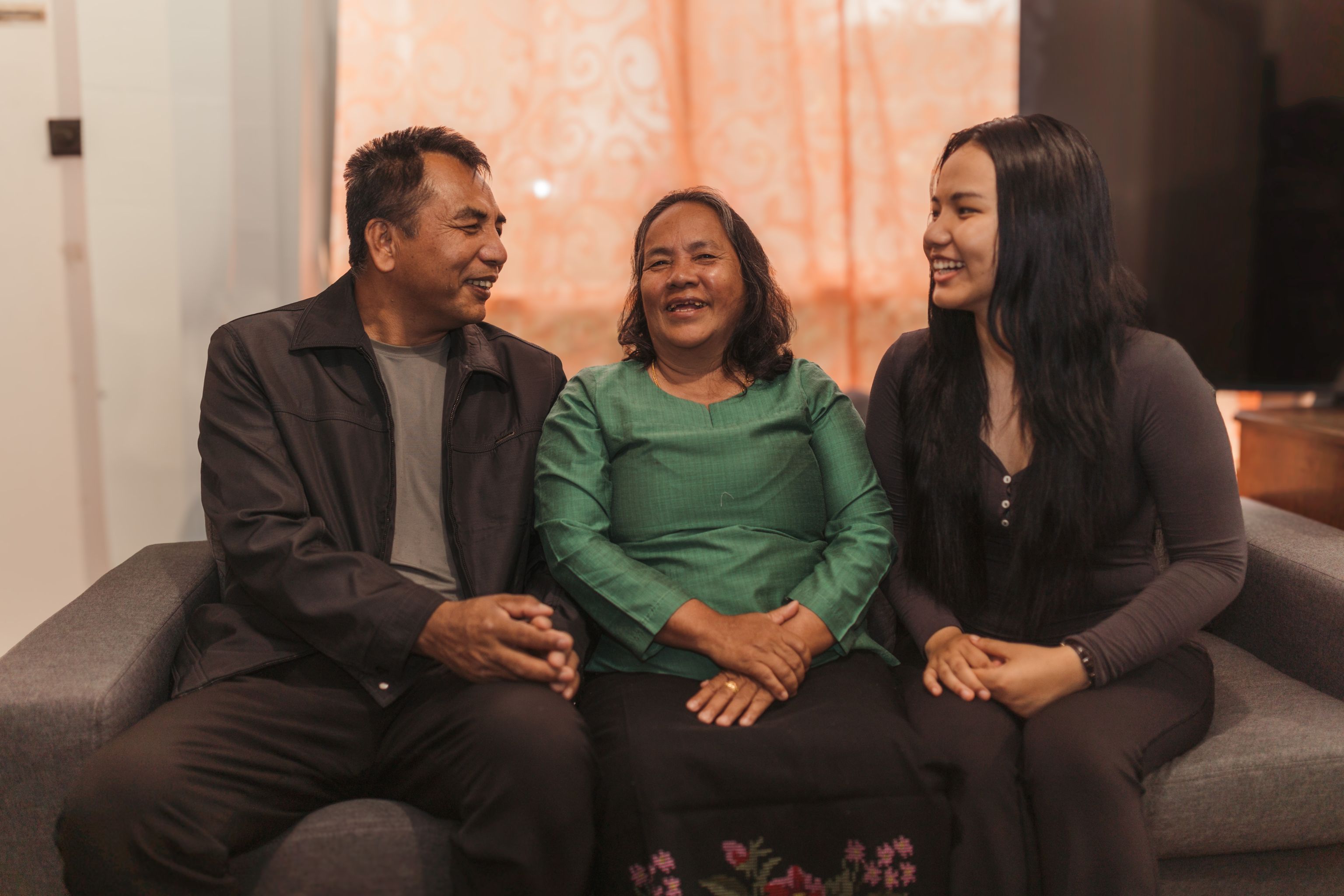 Father Van, mother Fung and daughter Daily sit together on a grey sofa, smiling happily to each other.