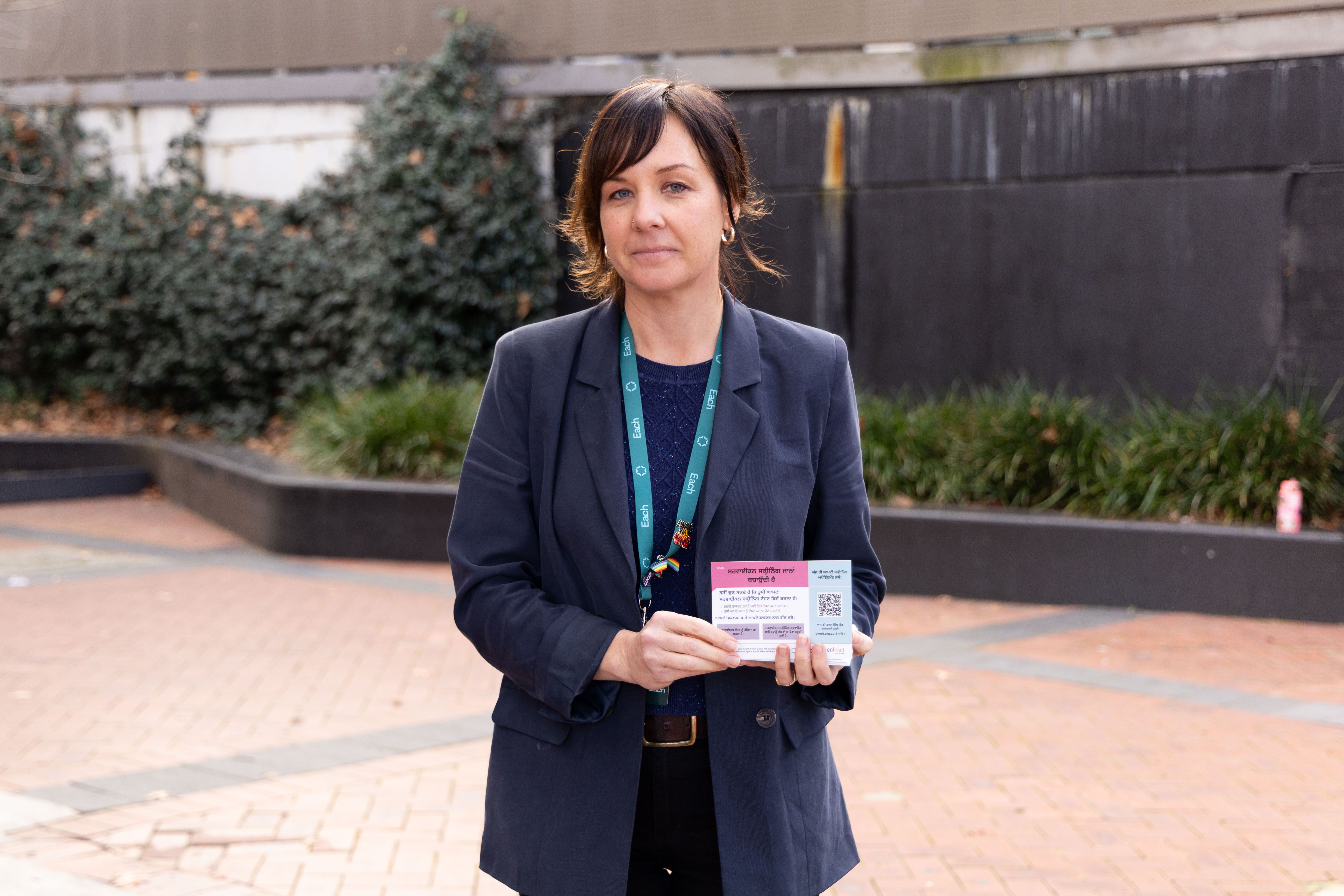 Rebecca, a young woman with dark hair waring a blue suit, is standing outside holding a card with information on it about cervical screening in Chinese