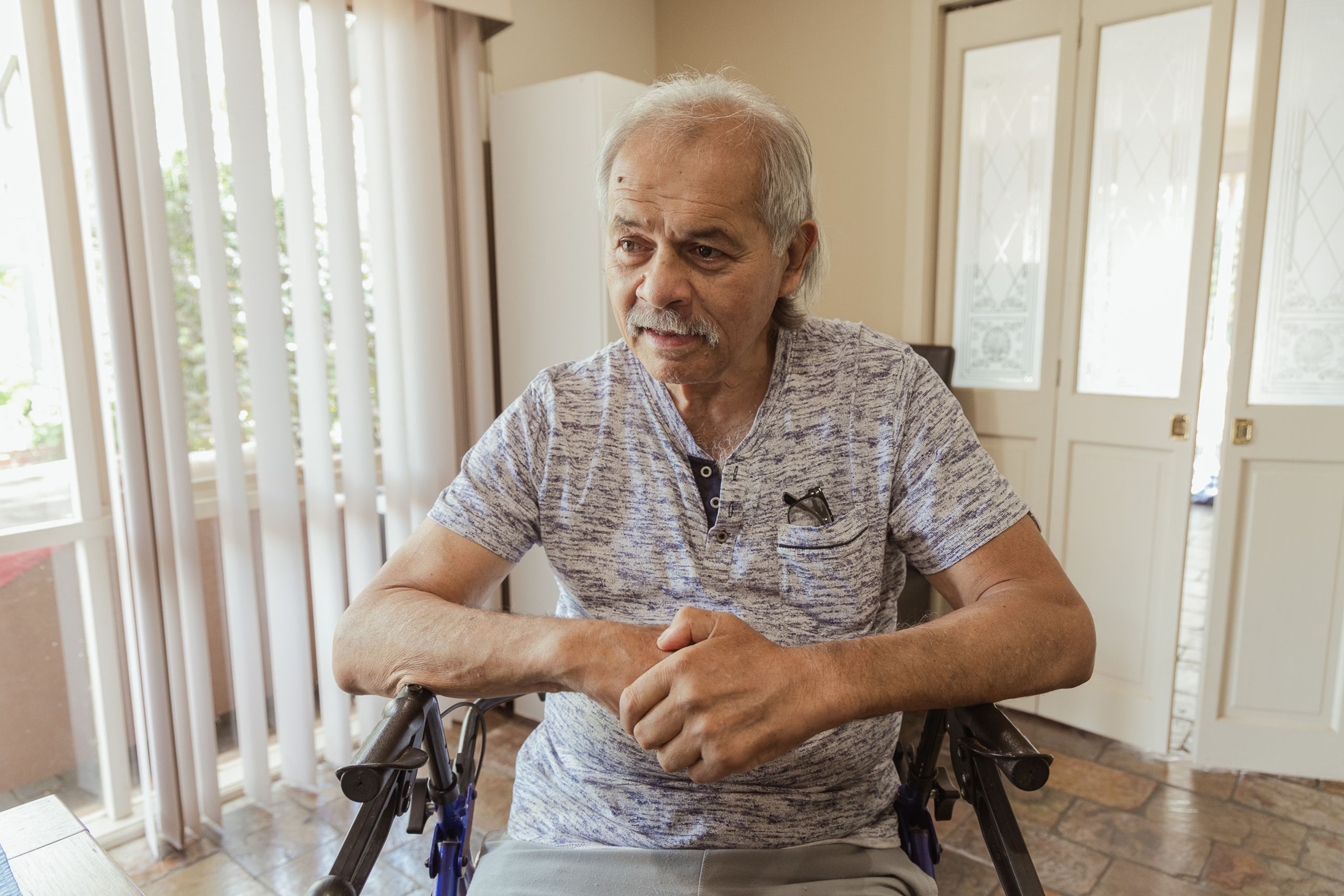An older man sits on a walker in his dining area, looking slightly to the side with a gentle smile.