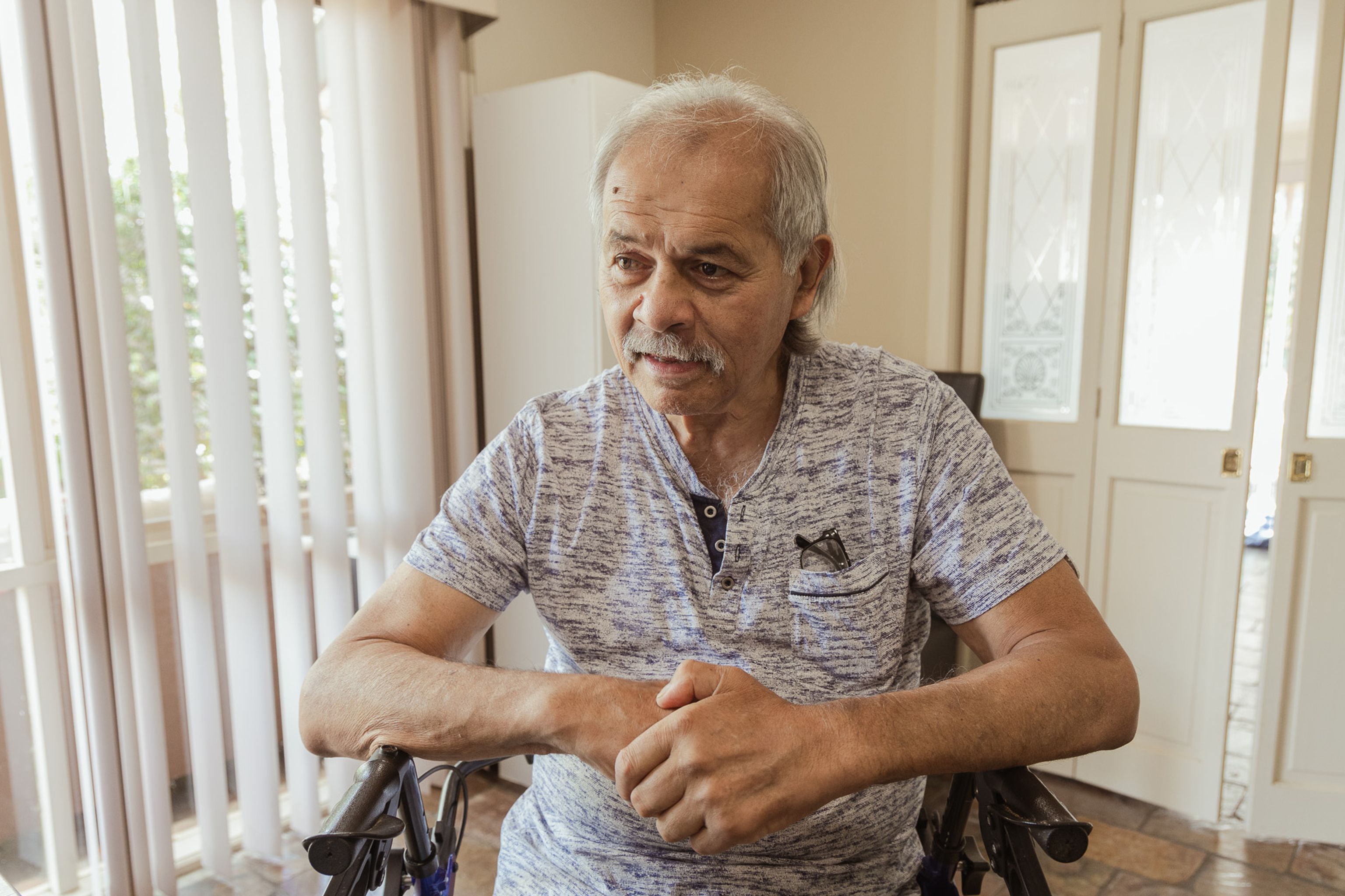 An older man sits on a walker in his dining area, looking slightly to the side with a gentle smile.