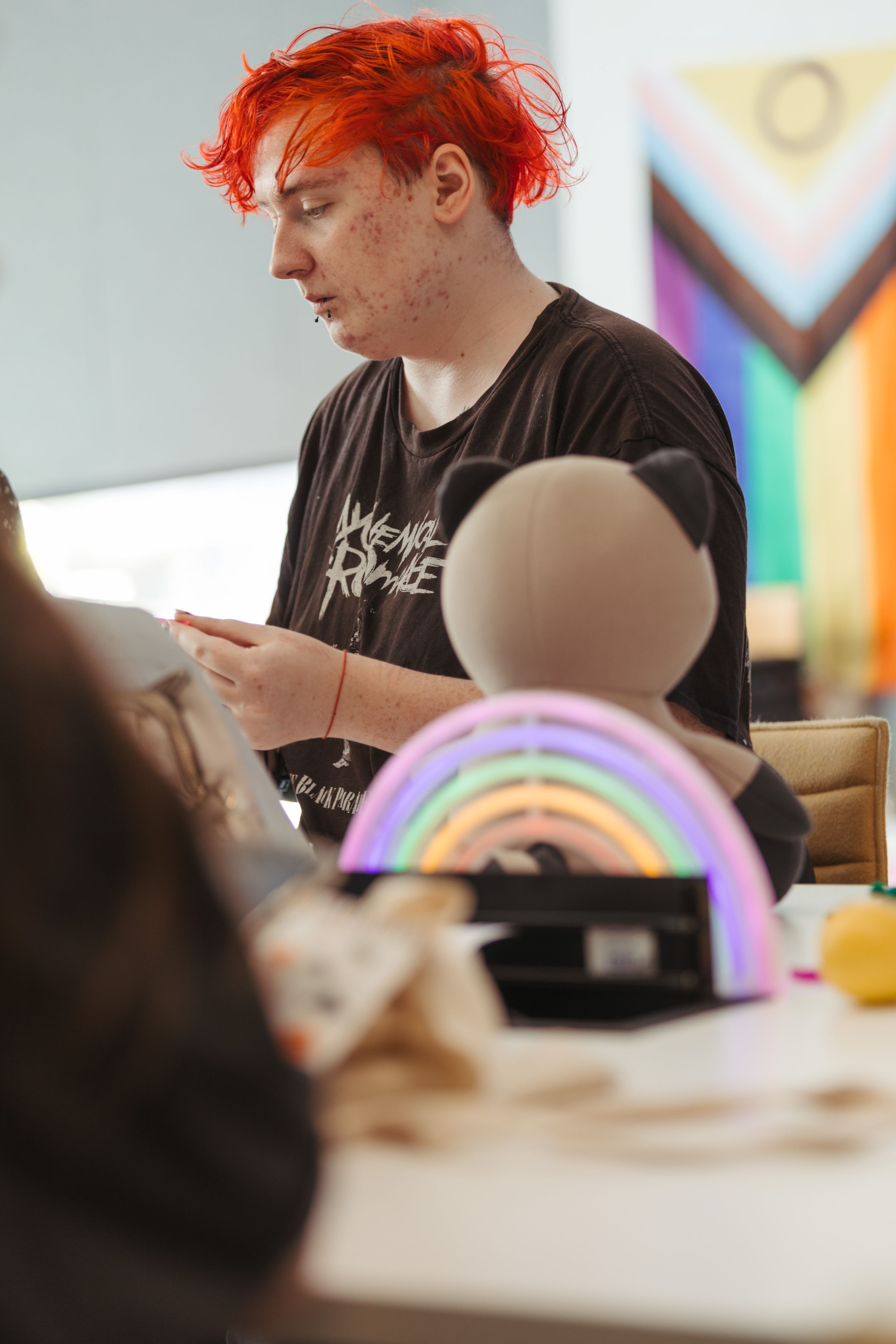 Nexus, a young adult, at a table. A rainbow flag in the background. A rainbow light and teddy bear are on the table.