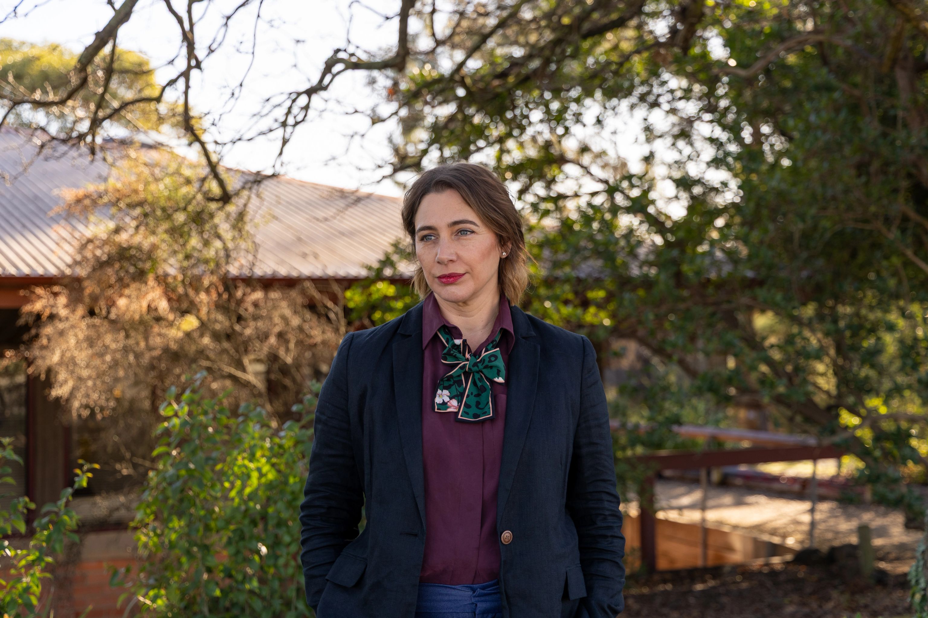 Kim Lucas stands in front of a clinic with trees behind her. She is looking to the left with her hands in her pockets and a slight smile.