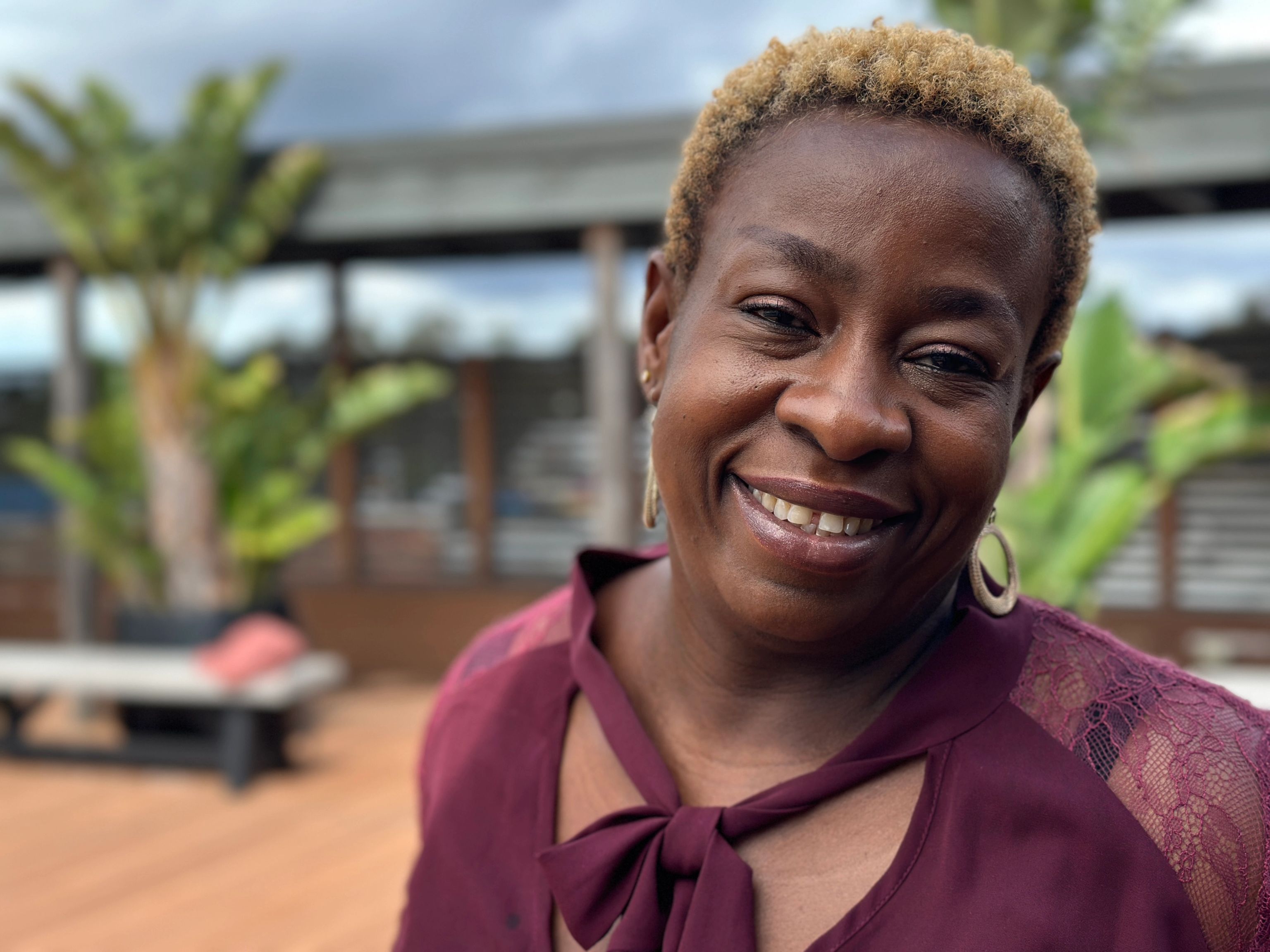 Person wearing a maroon top with a bow detail, standing outdoors near plants and wooden decking.