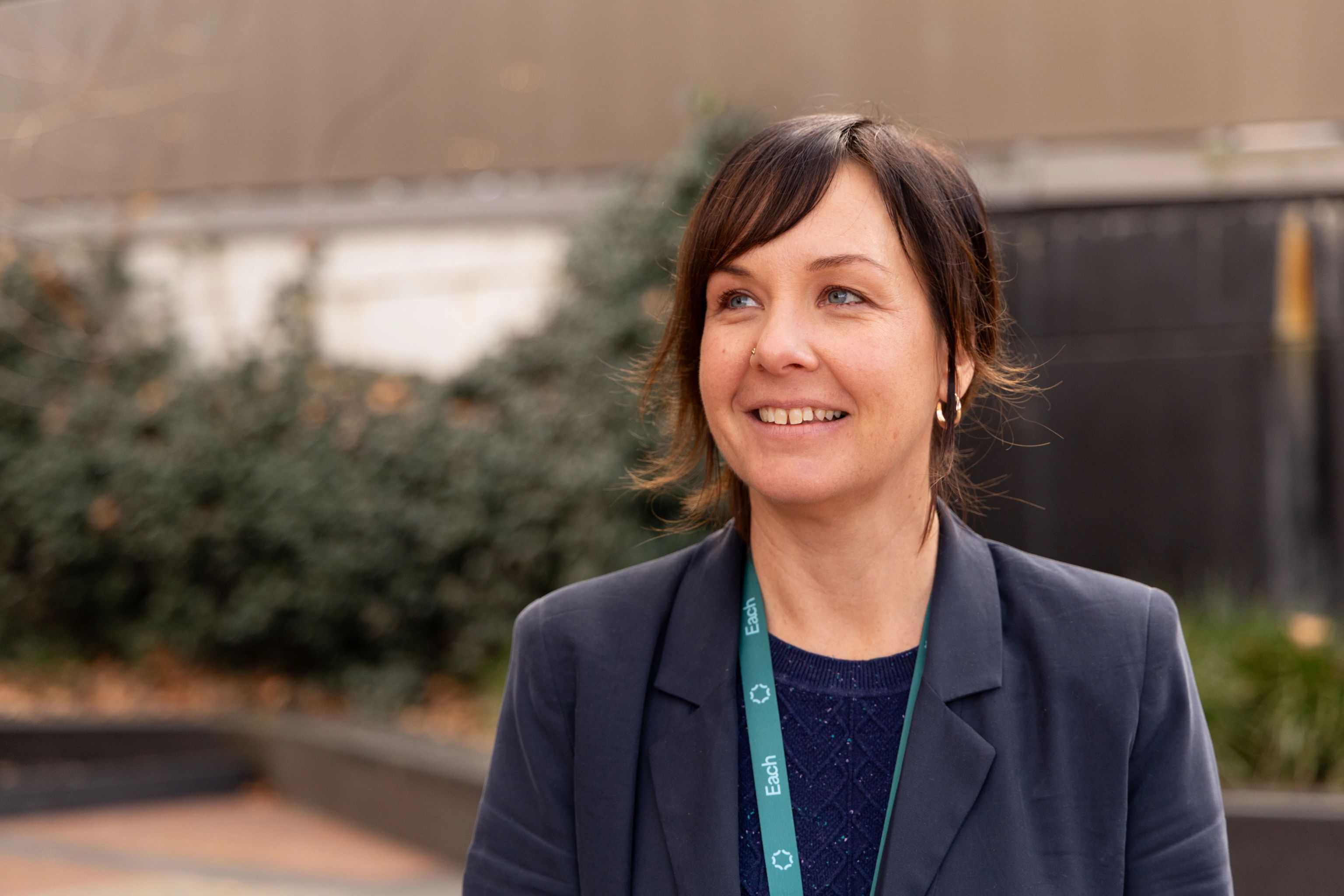Rebecca, a young women with dark hair, is standing outside in front of some greenery in a blue suit. She's smiling and looking off camera.