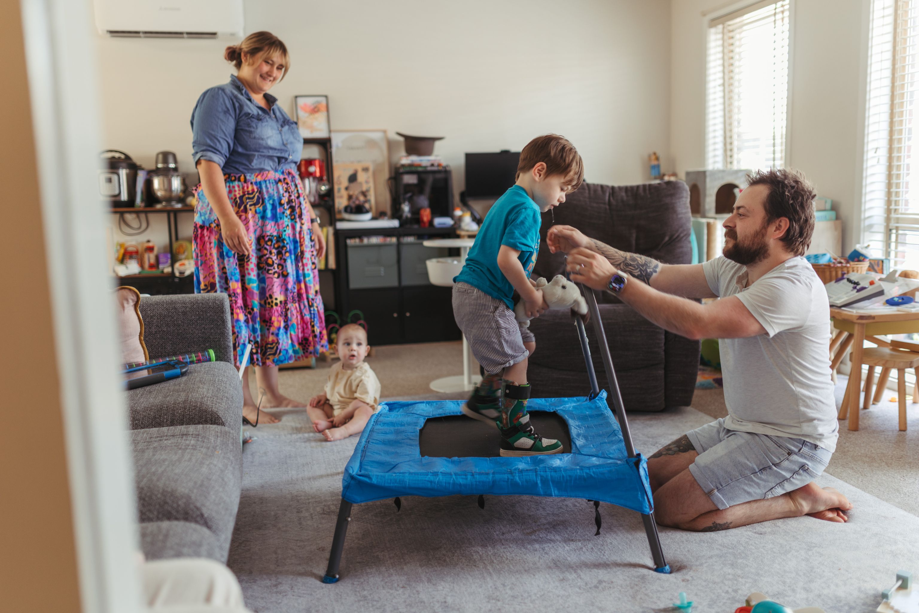 A happy family of 4 are together in a warm and busy living room. Four-year-old Henry is holding white bunny plushie while jumping on a small trampoline. His parents and baby sister watch him with big smiles.