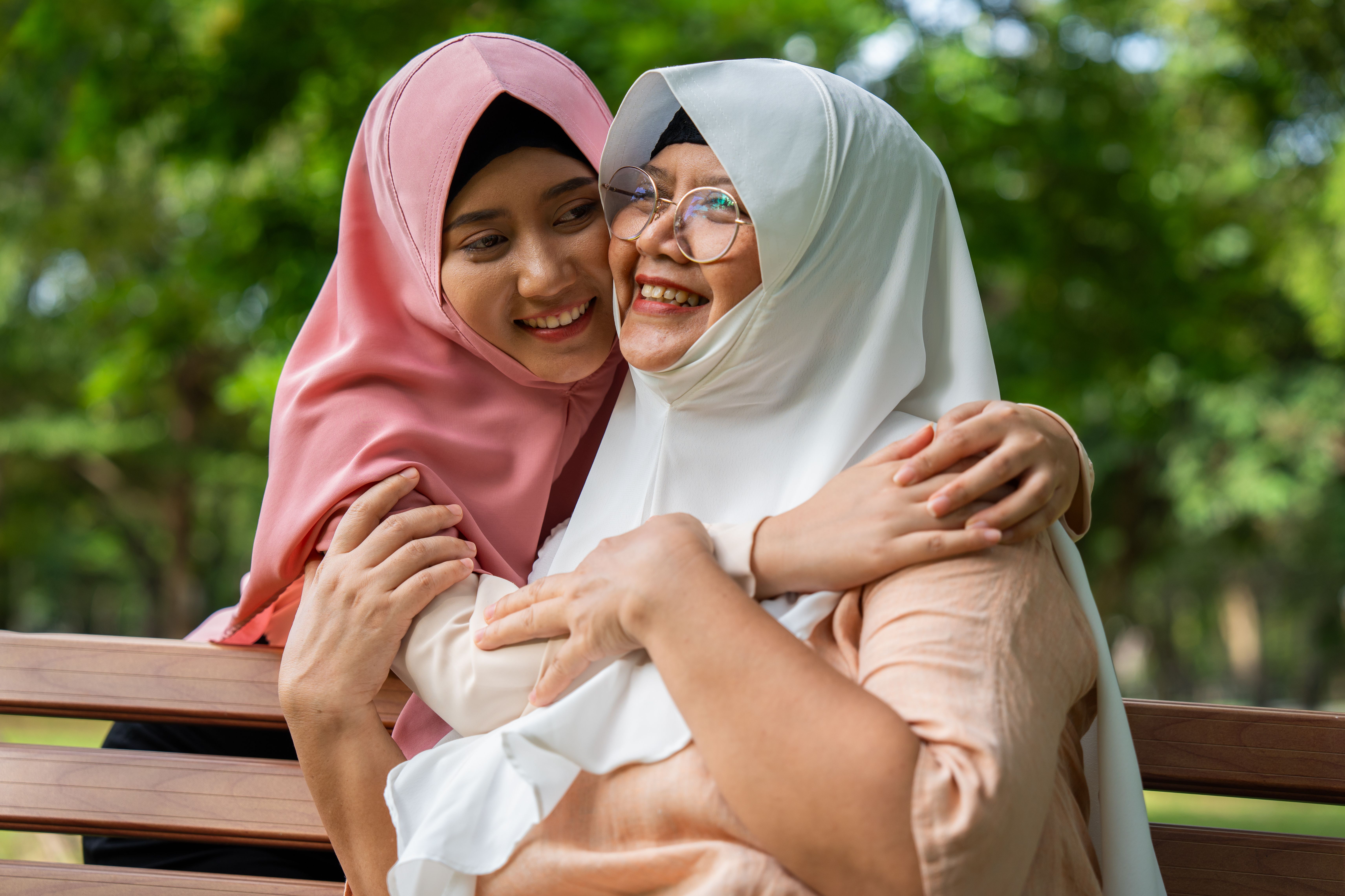 Two people wearing headscarves sit on a park bench, hugging each other outdoors.