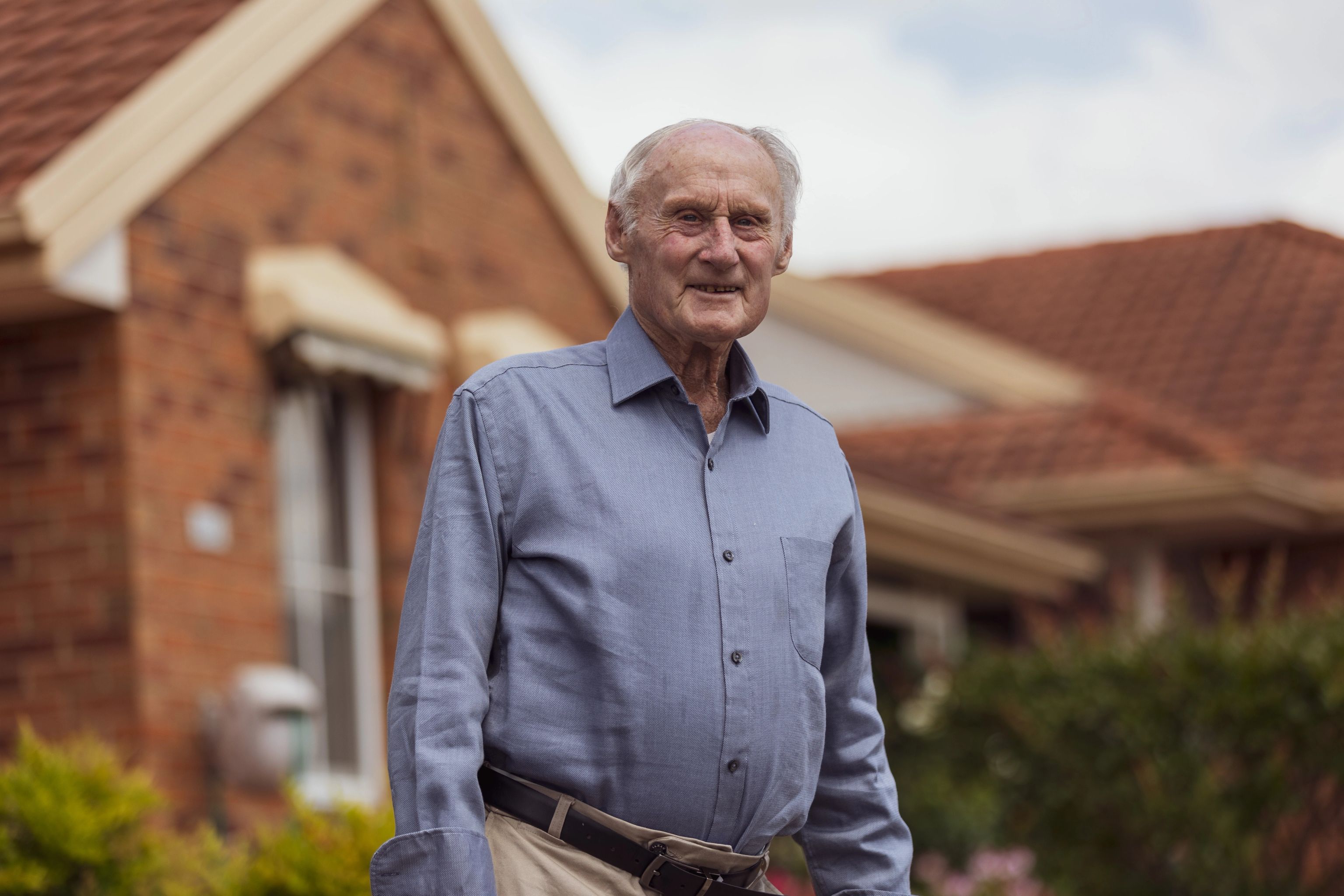 Older man wearing a blue button-up shirt and beige pants standing outside in front of a brick house with a tiled roof, smiling gently at the camera