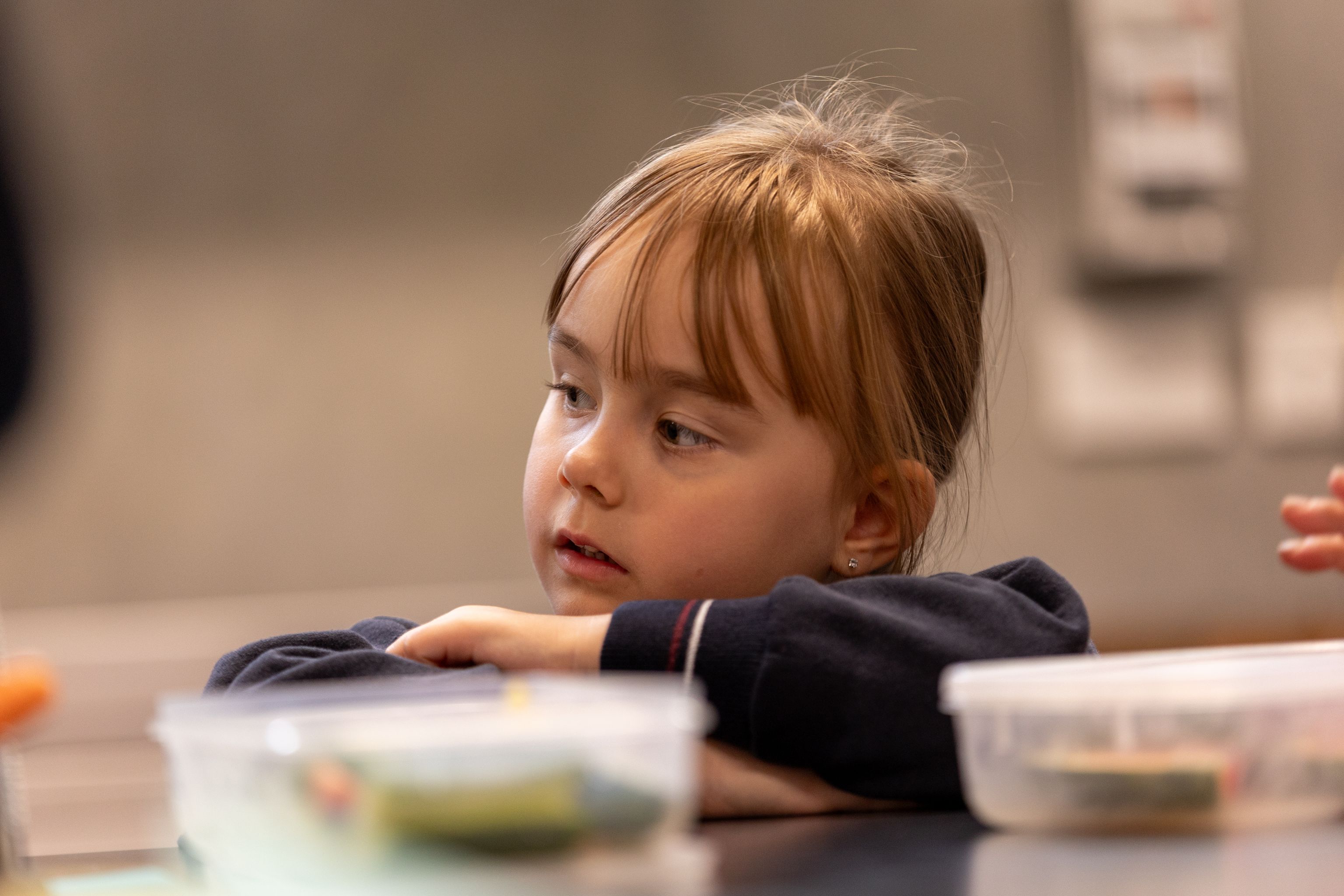 Close up of a 4 year old girl, resting her arms on a table and looking away to the distance, there are lunch boxes on the table in the foreground