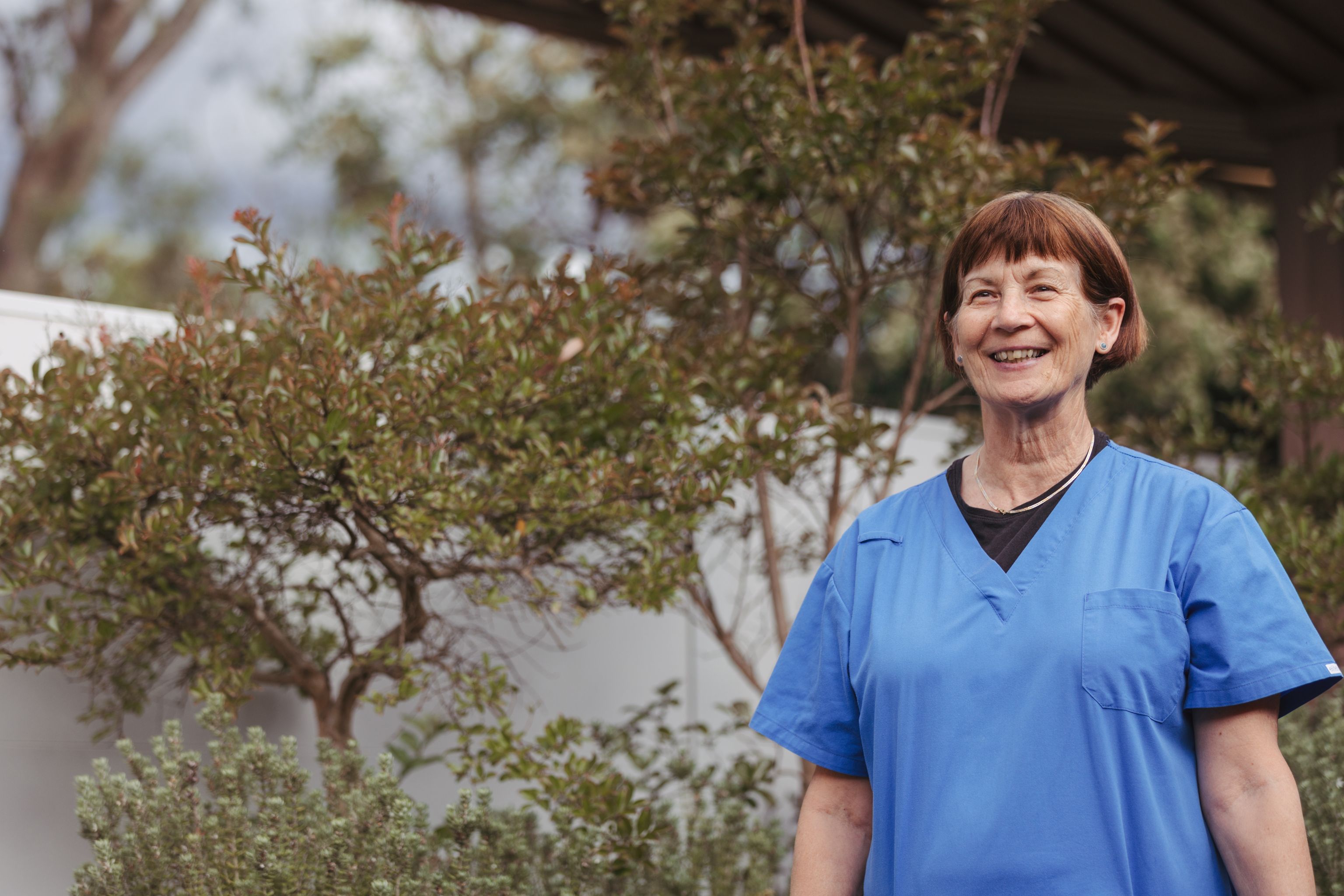 A middle aged Each staff worker wearing scrubs standing outside and smiling into the distance