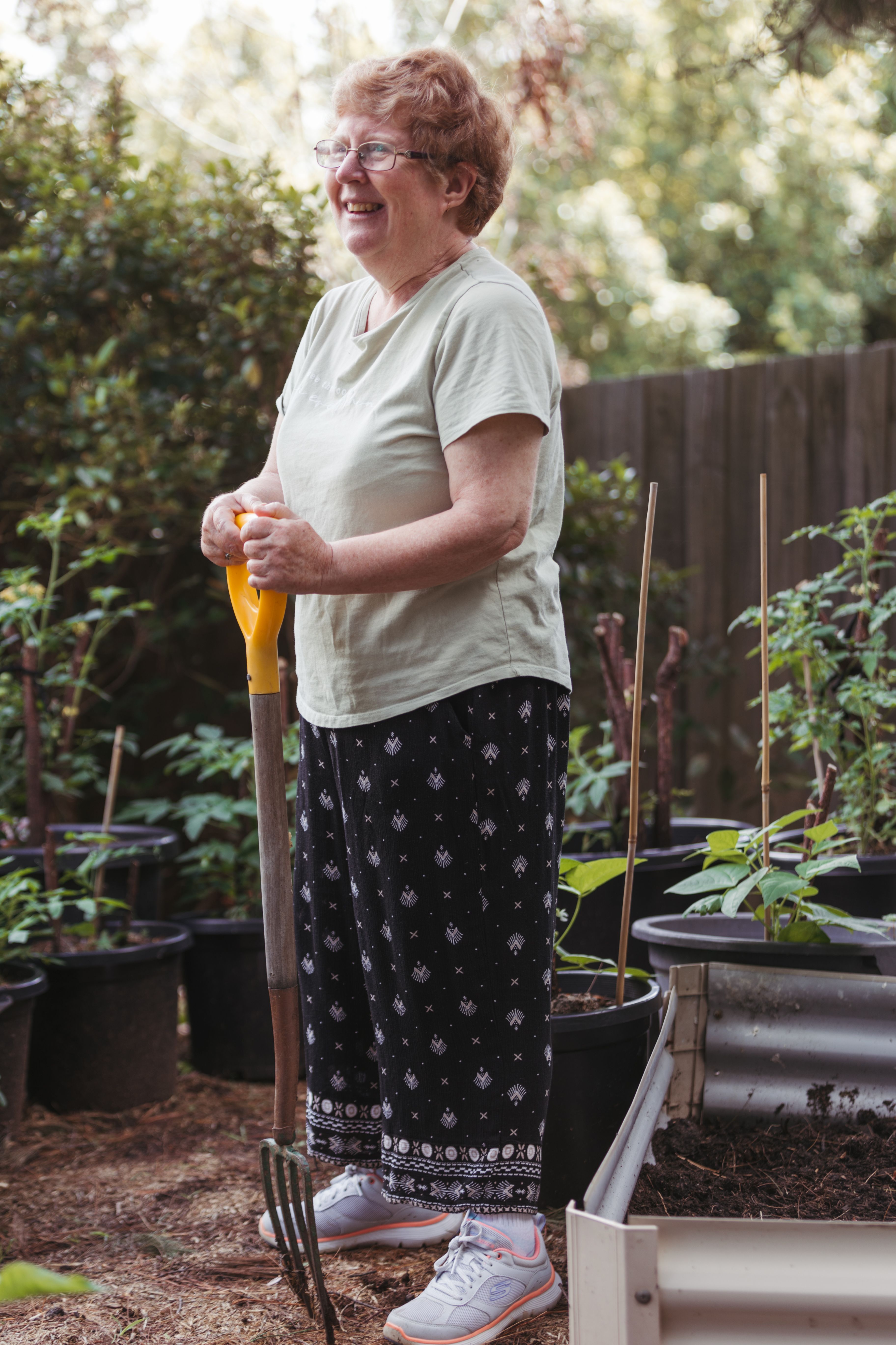 Senior woman standing in her garden, leaning on a pitch fork, and smiling