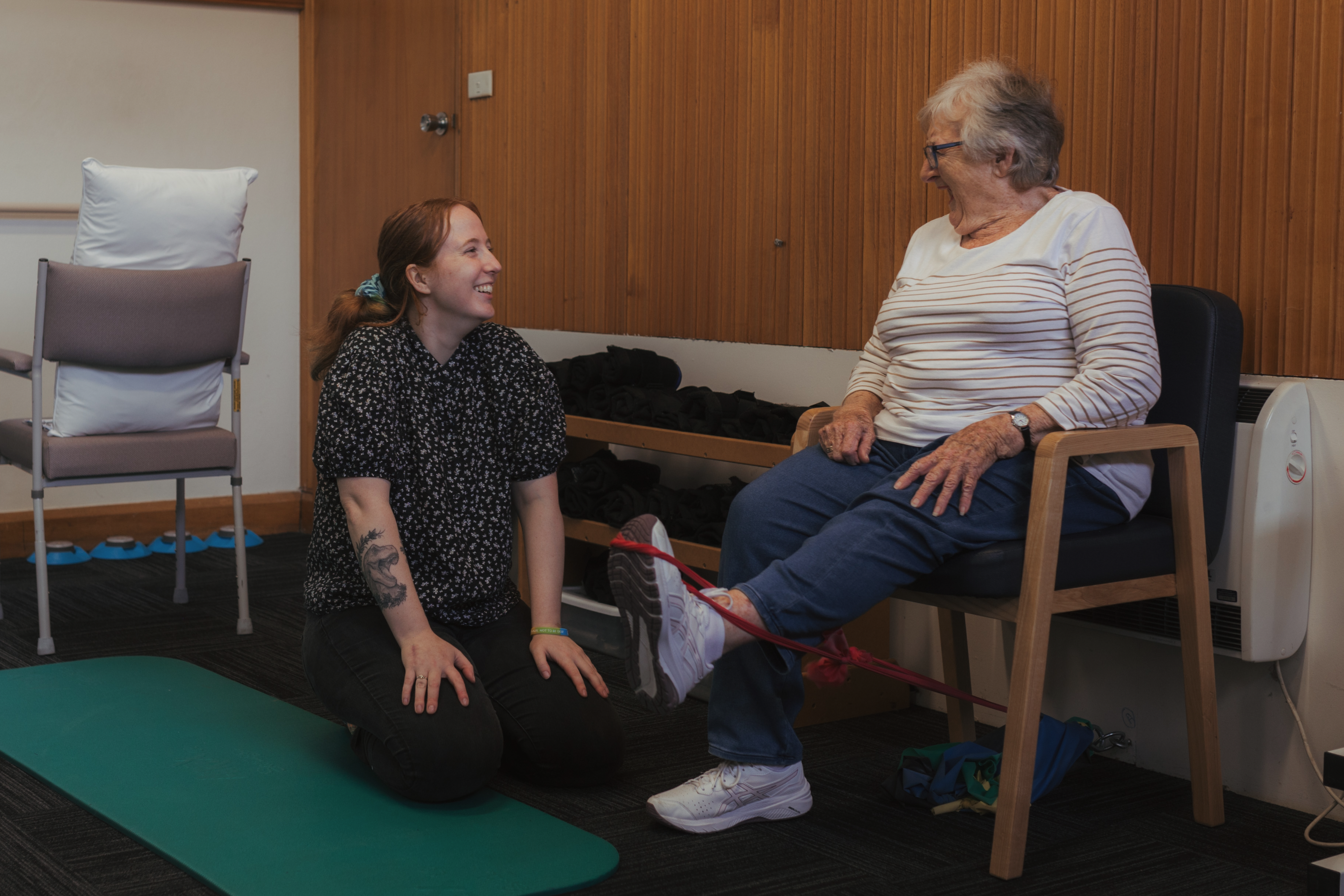 Older adult completing a seated resistance band exercise with support from a health professional in a community health setting.