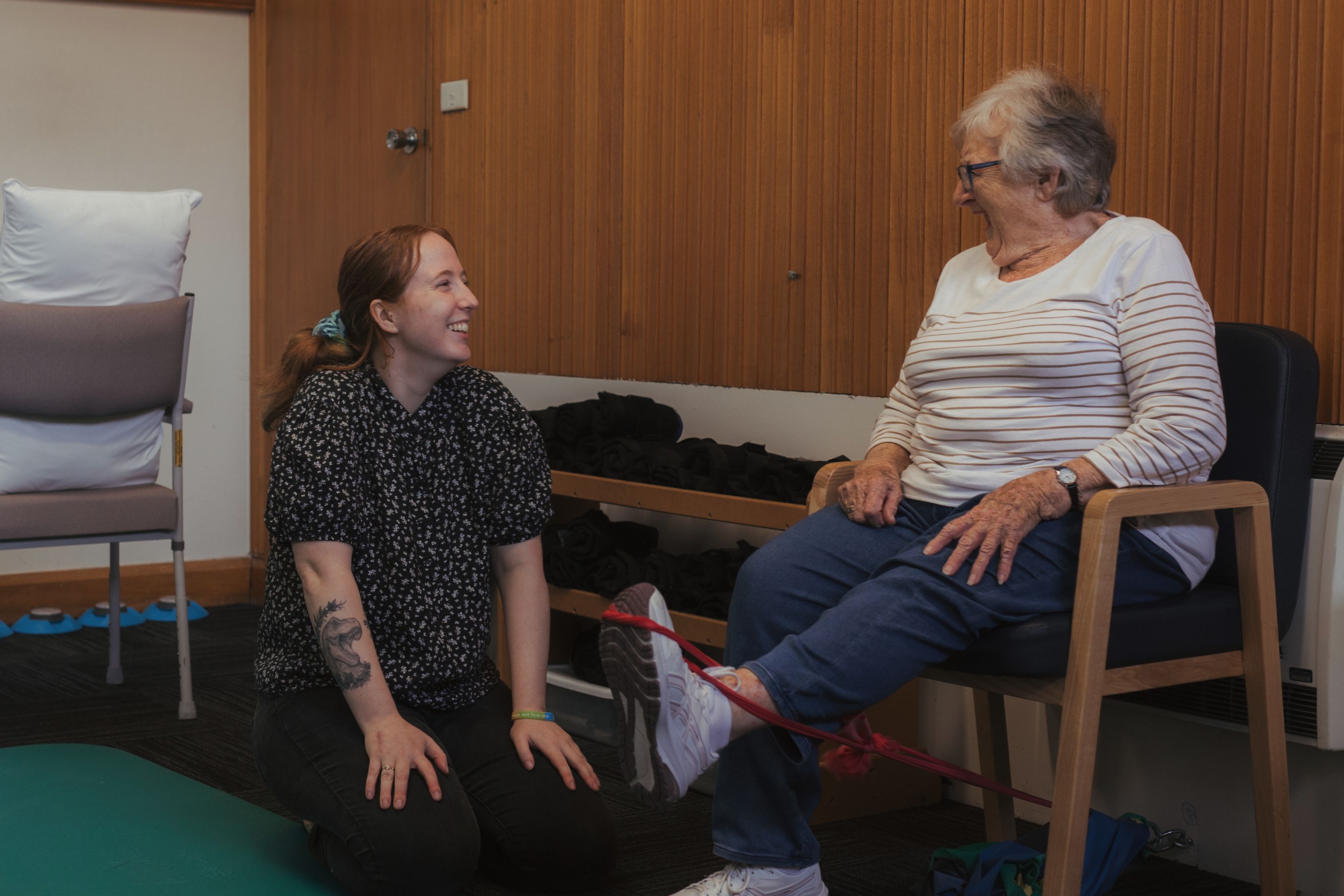 Older adult completing a seated resistance band exercise with support from a health professional in a community health setting.
