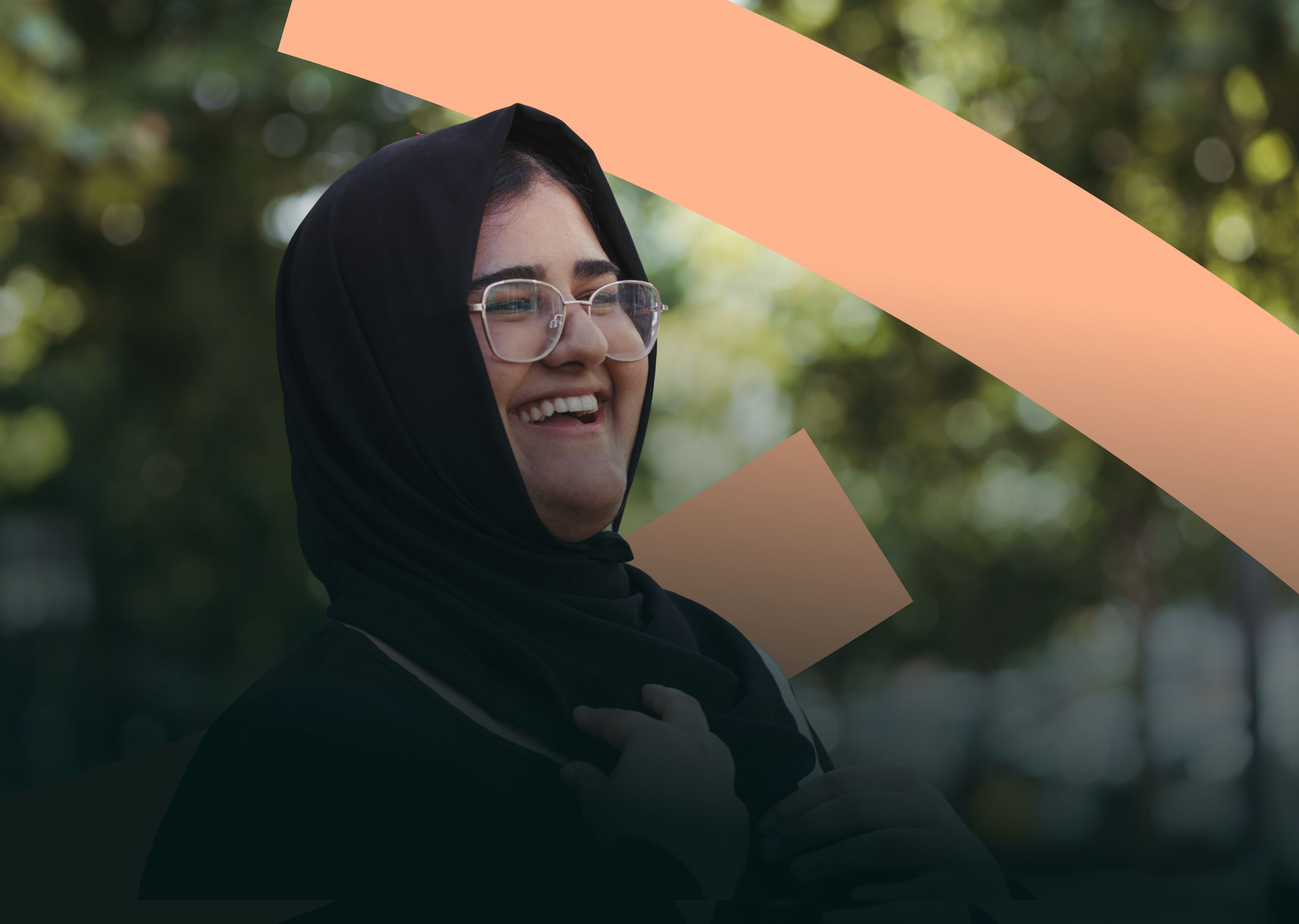 Close up of a woman wearing a black hijab smiling and looking into the distance, trees are in the distance