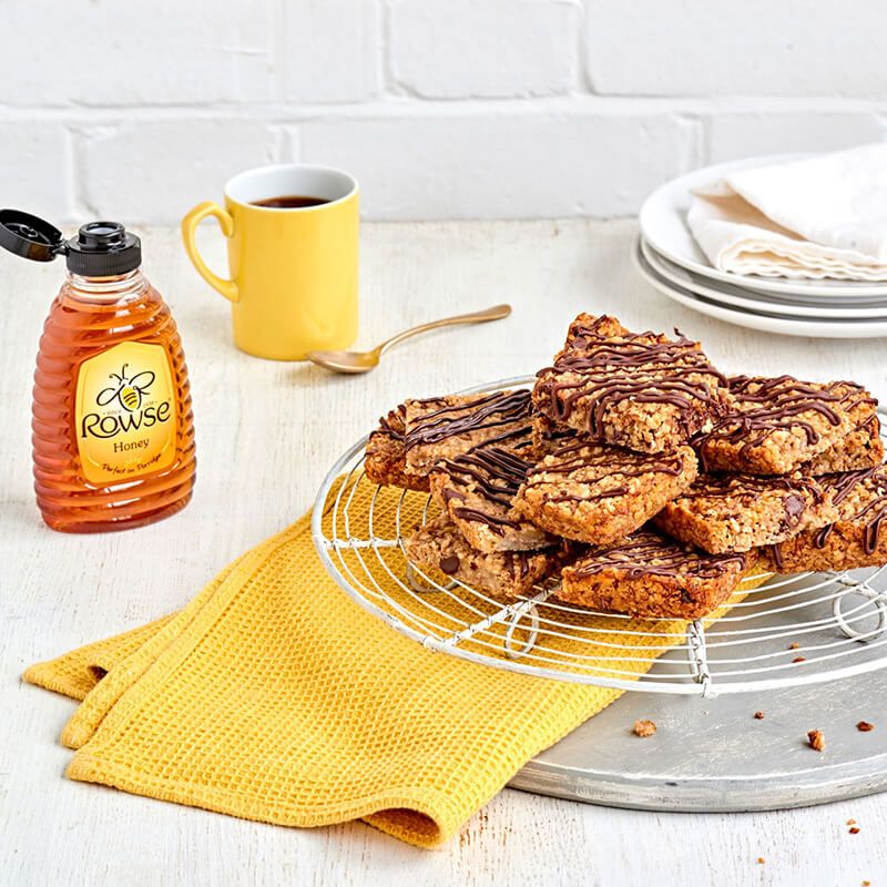 Rowse honey bottle, yellow coffee mug, and chocolate-drizzled oat bars on a wire rack.