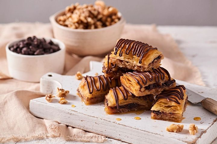 A white wooden chopping board with 6 chocolate drizzled slices of golden baklava and some drops of honey on the board, and a small bowl of dark chocolate chip and a bowl of walnuts in the background