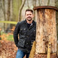 a man in a black jacket is standing next to a wooden structure in the woods .