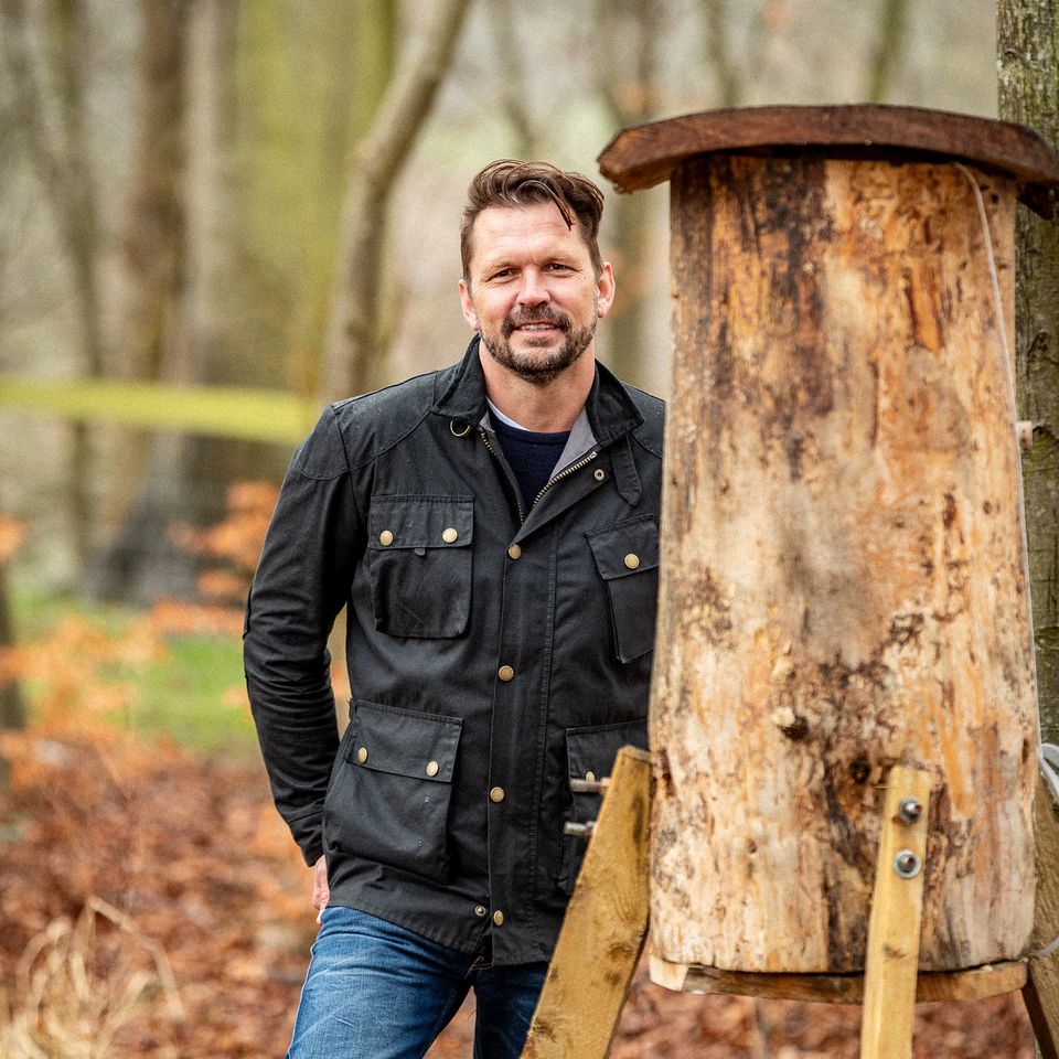 a man in a black jacket is standing next to a wooden structure in the woods .