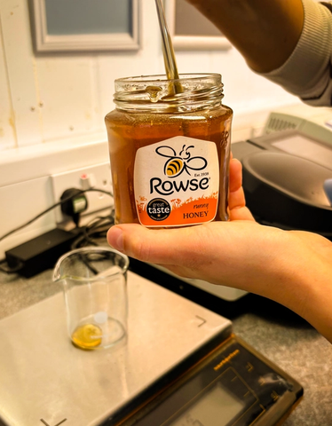 A person spoons honey from a Rowse honey jar above a digital scale with a beaker.