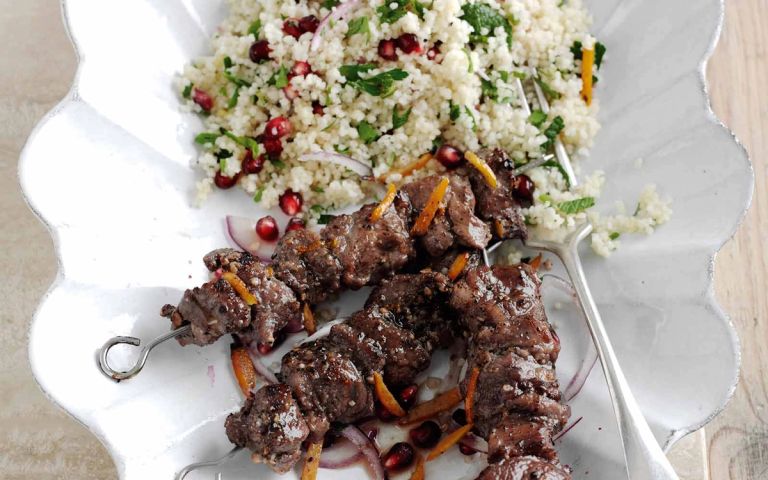 Two meat skewers garnished with orange zest and pomegranate, served with a couscous and herb salad on a white scalloped plate.