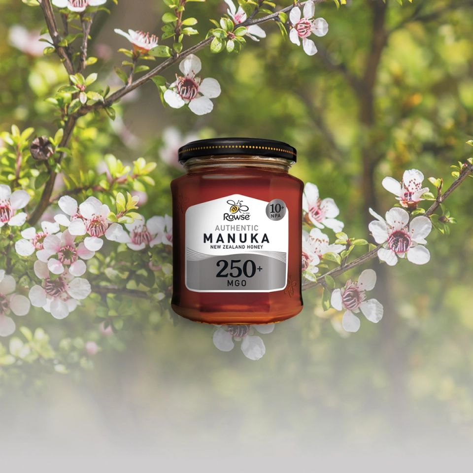 a jar of Rowse Manuka Honey 250+ in front of a tree with flowers .