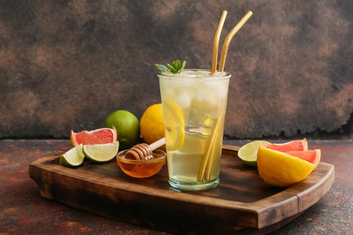 a glass of lemonade with straws , lemons , limes and honey on a wooden tray .