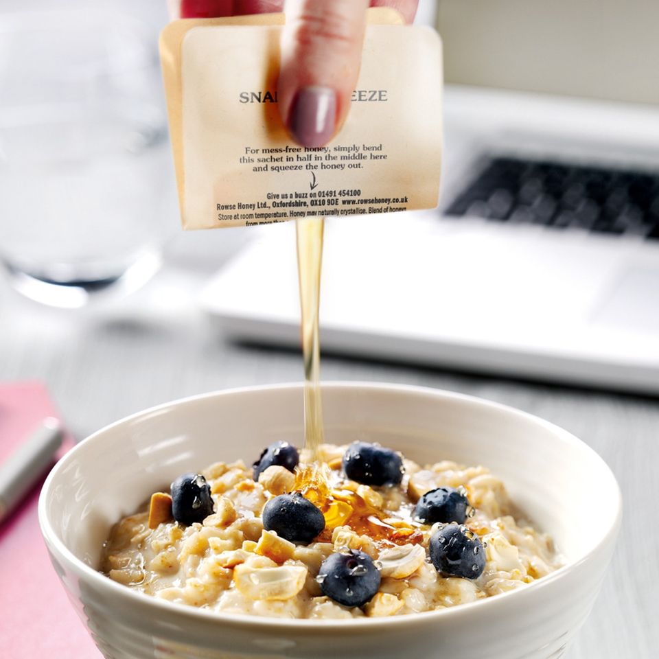 a person is pouring honey from Rowse Snap & Squeeze over a bowl of oatmeal with blueberries .