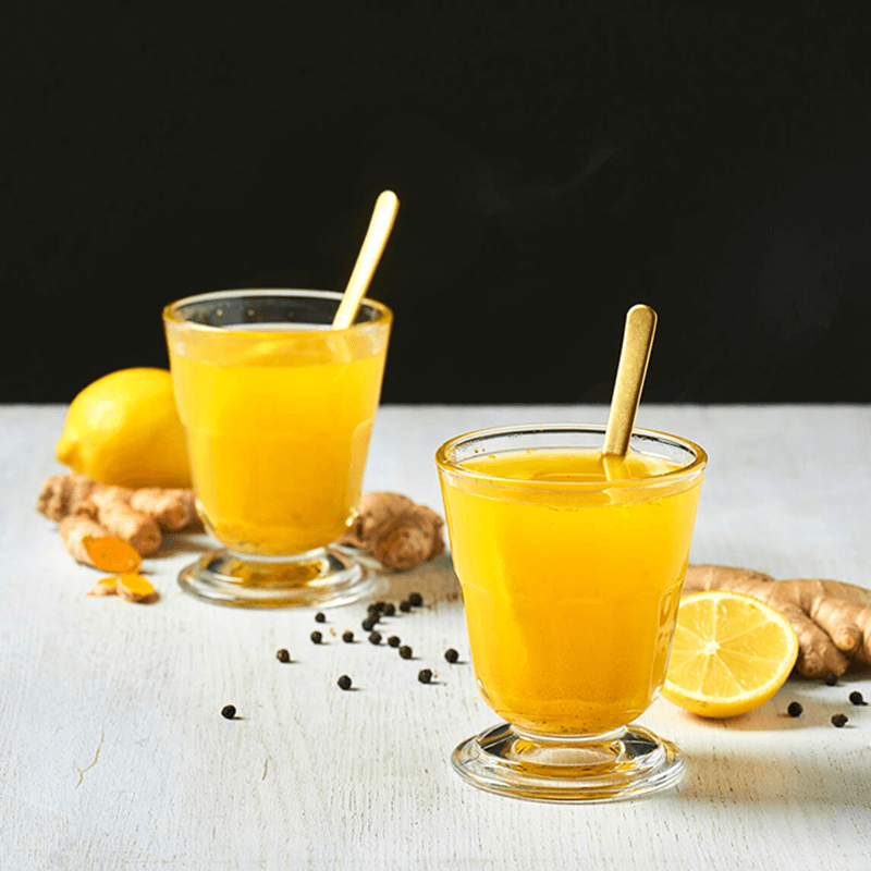 Two glasses of golden drink with gold spoons, surrounded by ginger, lemon, turmeric, and peppercorns.