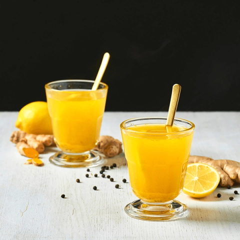 Two glasses of golden drink with gold spoons, surrounded by ginger, lemon, turmeric, and peppercorns.