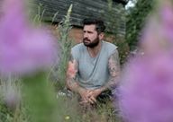 a man with a beard and tattoos is sitting in the grass surrounded by purple flowers .