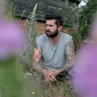 a man with a beard and tattoos is sitting in the grass surrounded by purple flowers .