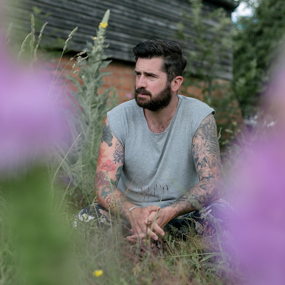 a man with a beard and tattoos is sitting in the grass surrounded by purple flowers .