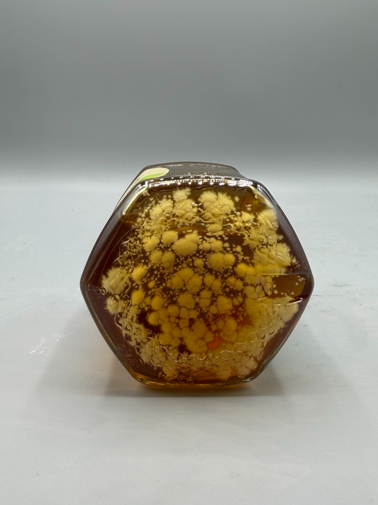 A hexagonal jar of dark amber honey with yellow mold growing inside.
