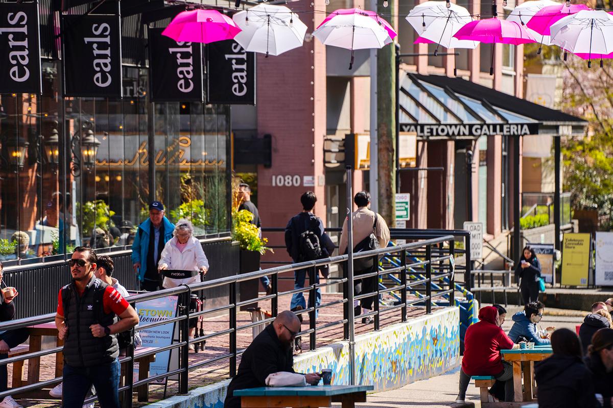 A busy outdoor area in Canada with people walking and sitting at picnic tables, colorful pink and white umbrellas hanging overhead, and a restaurant named 'Earls' in the background.
