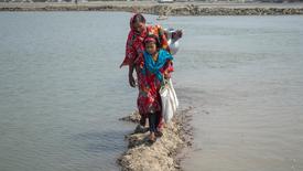 A woman and a young girl in colorful clothing walking along a narrow path surrounded by water in Bangladesh. The woman is carrying a metal pot on her shoulder.