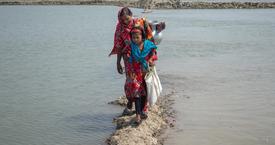 A woman and a young girl in colorful clothing walking along a narrow path surrounded by water in Bangladesh. The woman is carrying a metal pot on her shoulder.